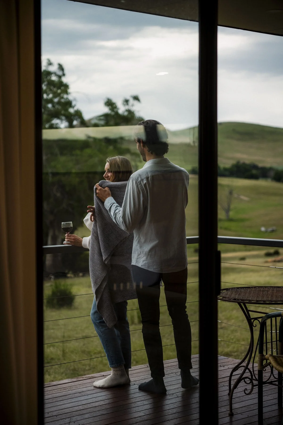 A couple enjoying a quiet moment on a balcony, with one woman holding a glass of red wine, while the man wraps a blanket around her shoulders, overlooking a scenic countryside landscape.