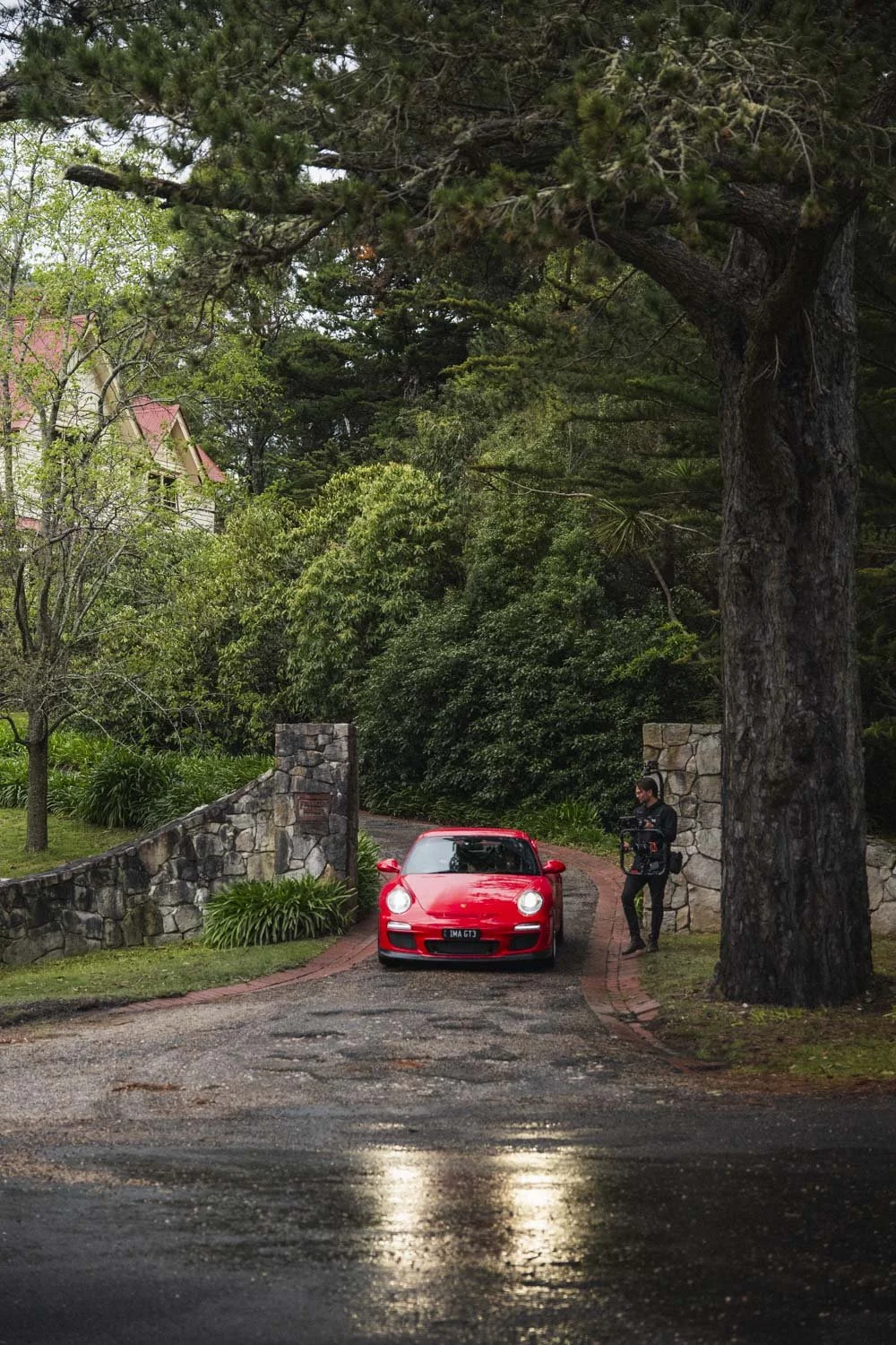 A red sports car parked on a curved driveway next to a person holding a camera or filming equipment, surrounded by large trees and a stone wall entrance gate, with a house partially visible in the background.