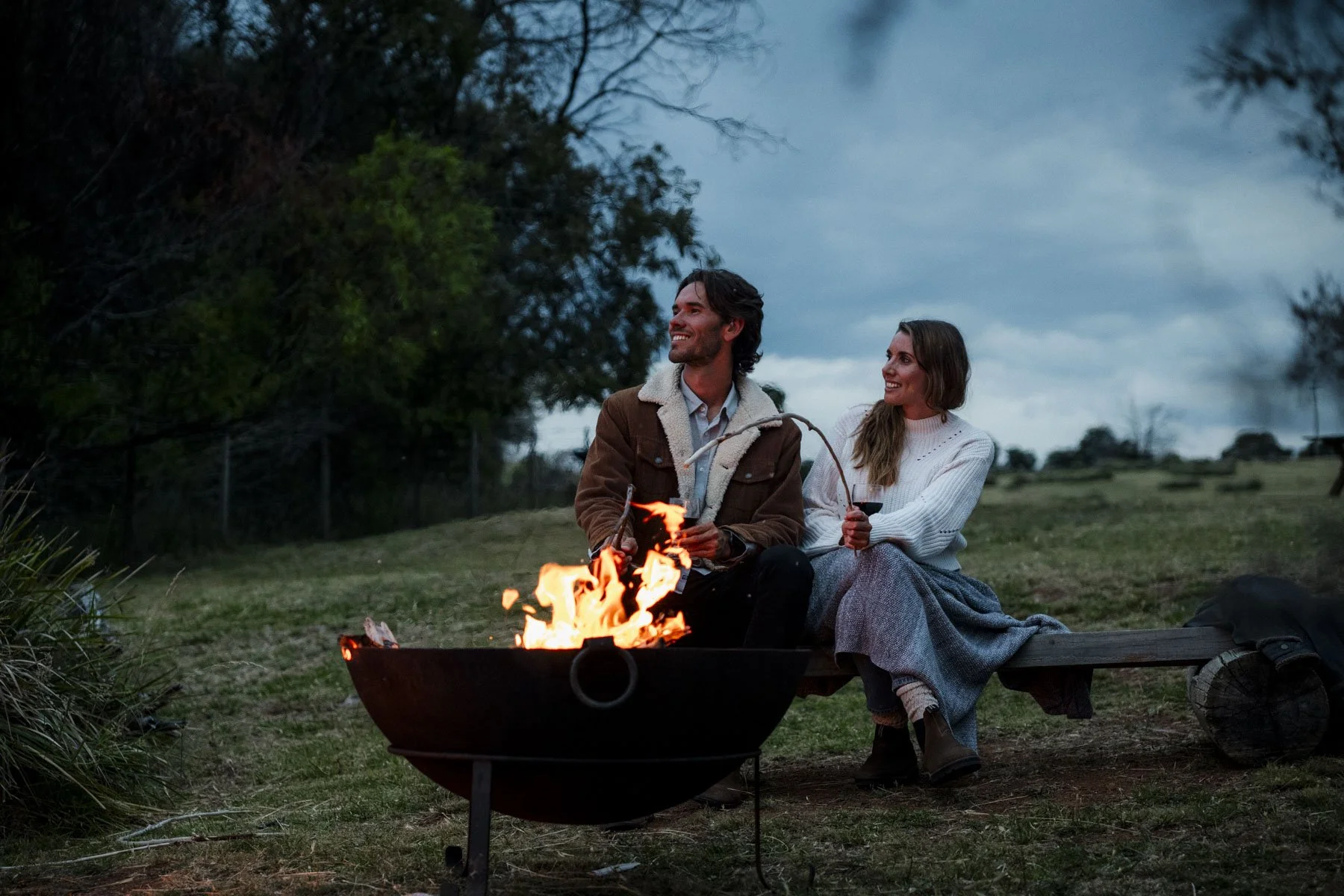 A man and woman sitting by a campfire on a wooden bench outdoors during the evening, sharing drinks and smiling at each other with trees and a cloudy sky in the background.
