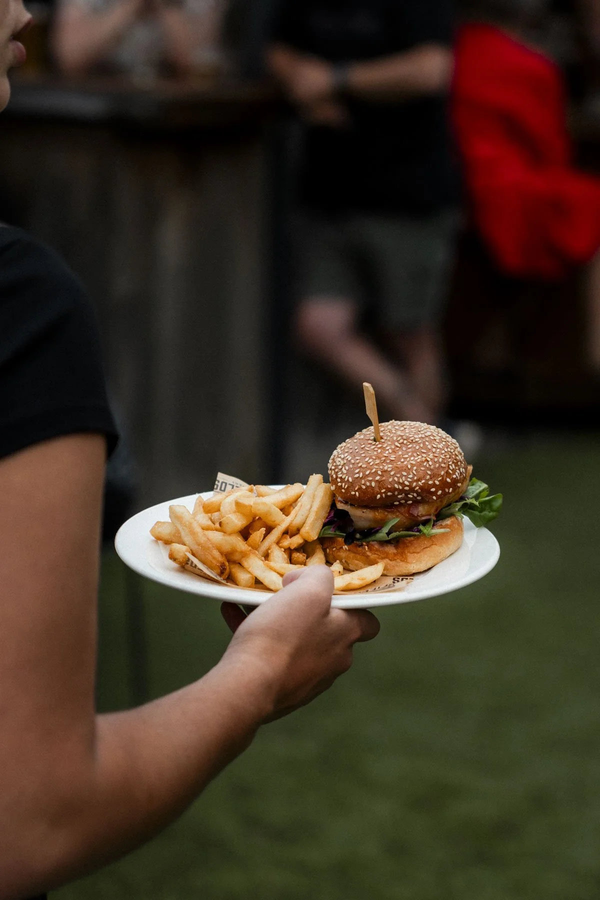Person holding a white plate with a cheeseburger and French fries, at an outdoor event with people in the background.