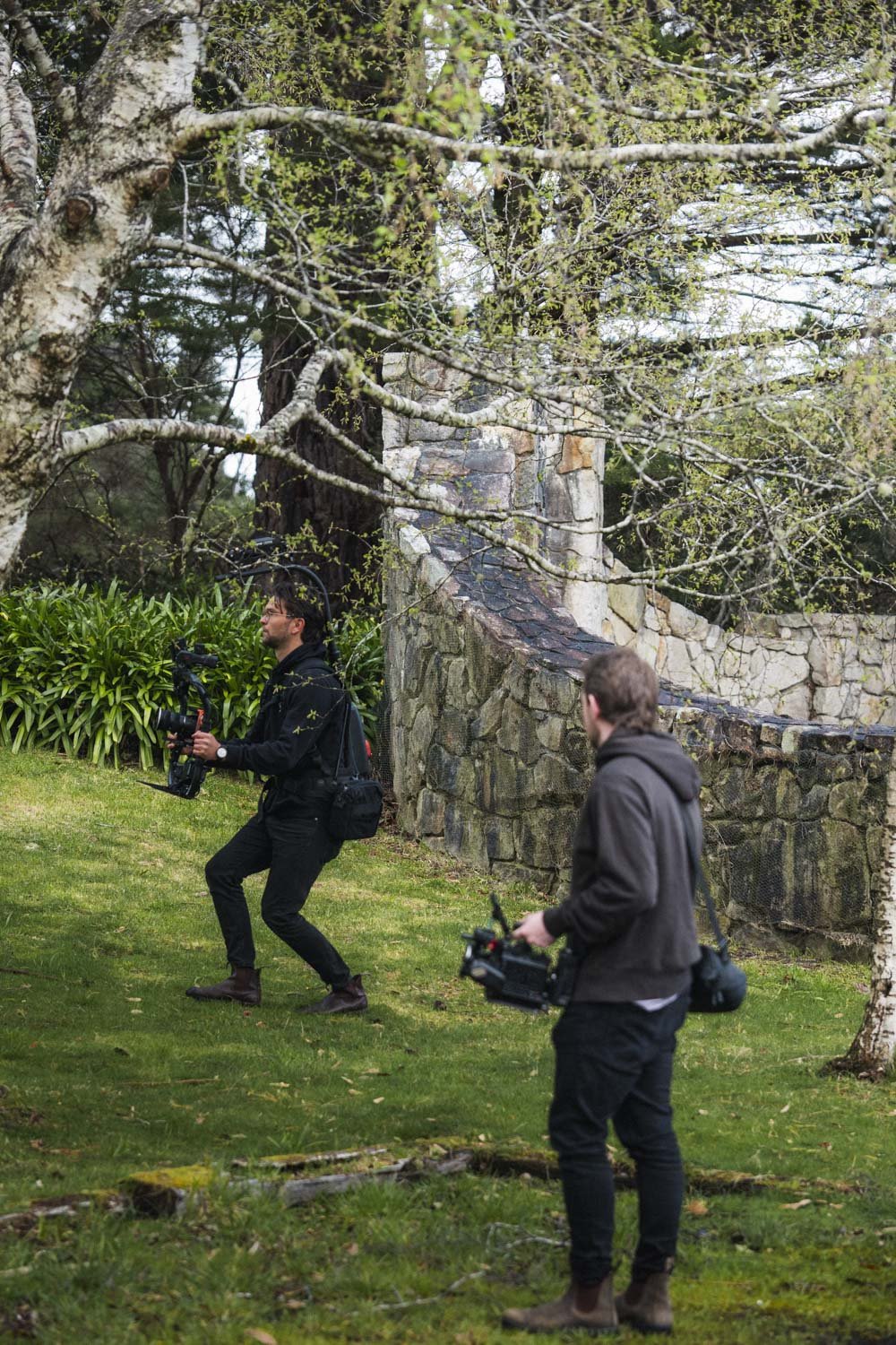Two men filming outdoors in a park-like setting, surrounded by trees and greenery.