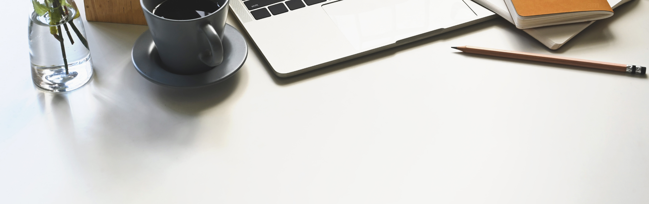 Minimal workspace with a silver laptop partially open beside a black coffee mug on a matching saucer. A pencil and stacked notebooks rest nearby with a small glass vase holding green stems, suggesting a calm and organized work setup.