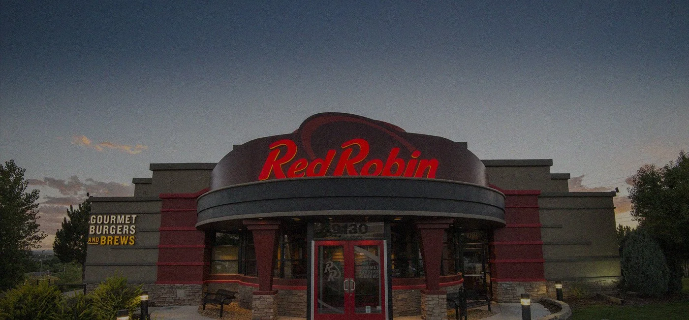 Exterior of a Red Robin restaurant with the sign and entrance visible at dusk, surrounded by small plants and trees.