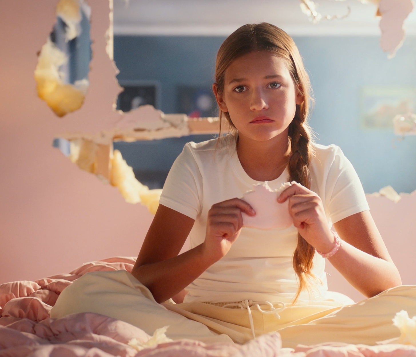 A teenage girl with long hair in a braid sitting on a bed, looking sad or upset, holding a torn piece of paper or tissue, with a large hole in a wall behind her.