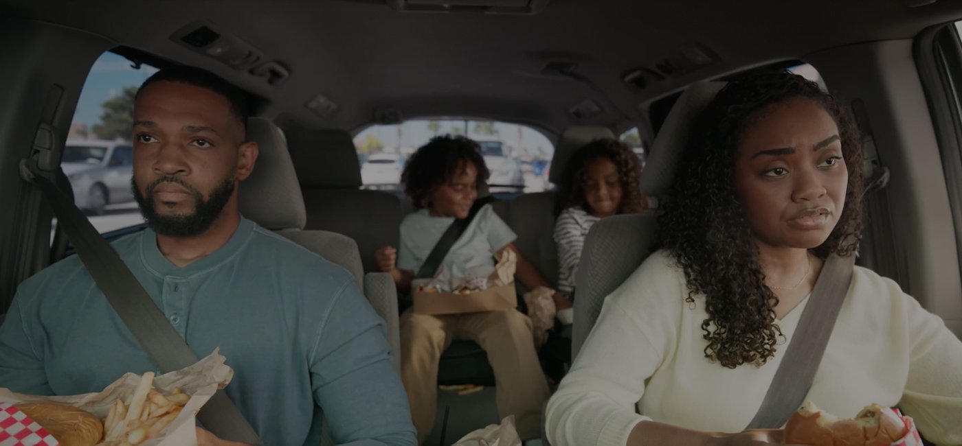 A family inside a car, with adults in the front seats and children in the back, eating fast food during a car trip.