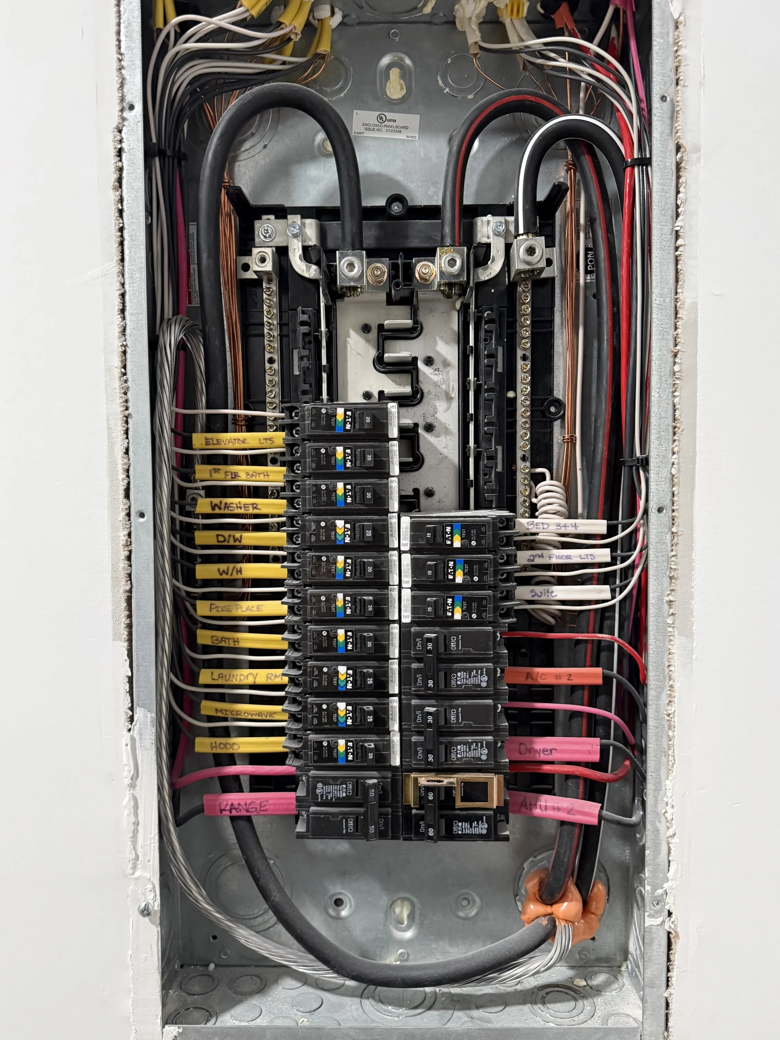 Electrical panel with various circuit breakers, wires, and yellow, pink labels indicating different circuits such as elevator, bathroom, washer, and microwave.
