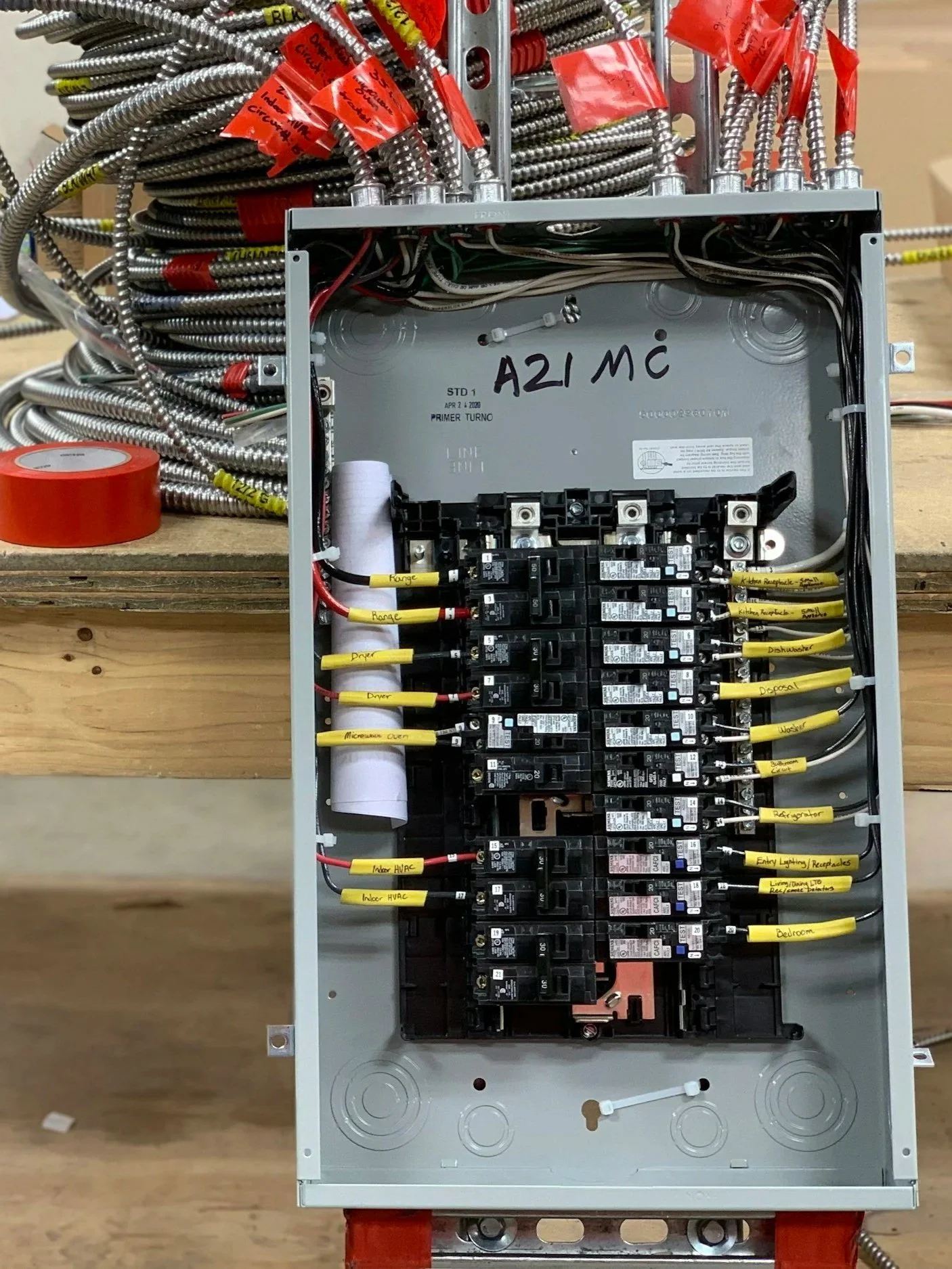 An electrical breaker panel with multiple labeled yellow wires connected to circuit breakers. The panel is open with bundles of metal conduit and red tape nearby.