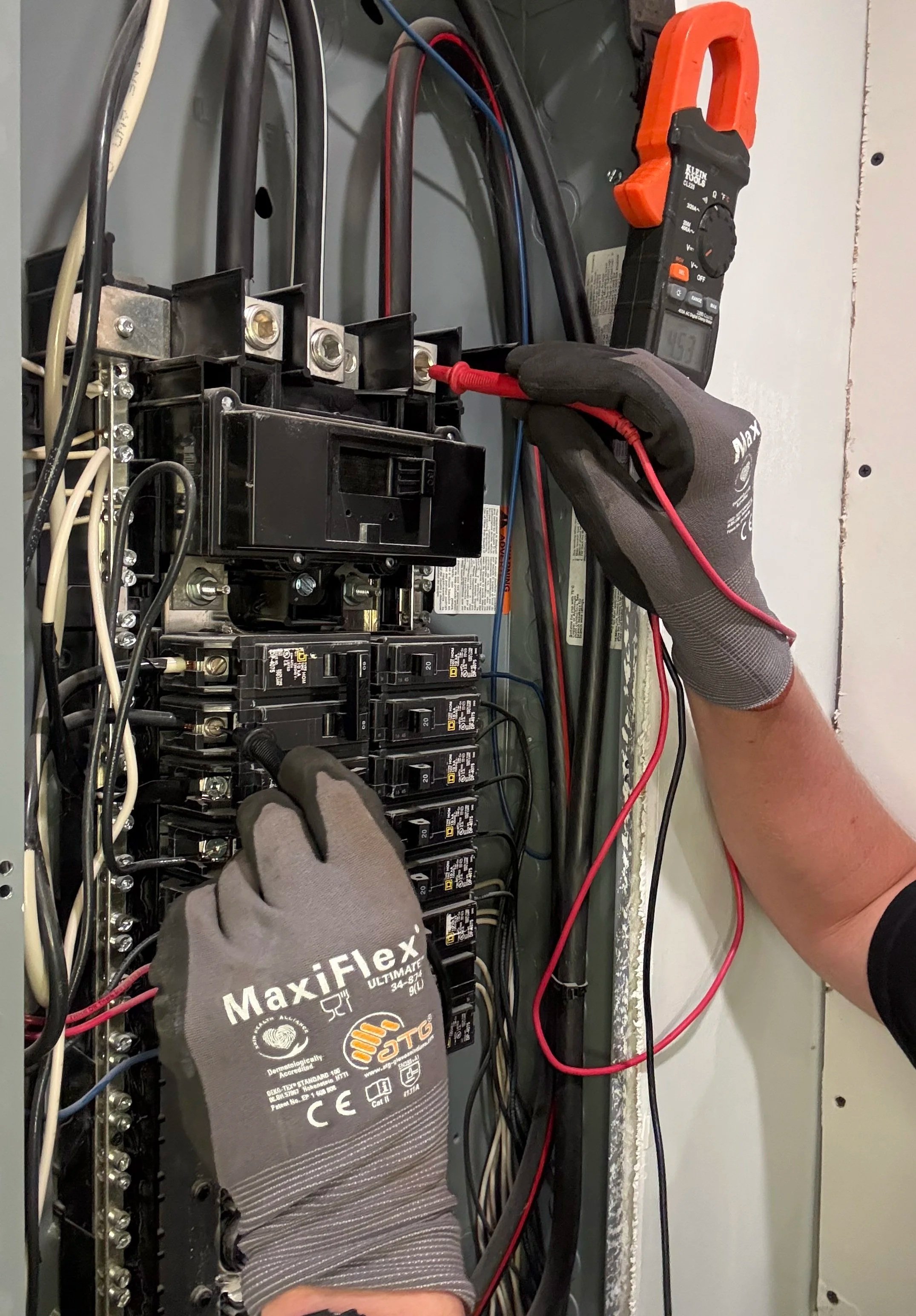 A person wearing gray Maxiflex gloves is using a multimeter to test electrical connections inside an electrical panel or breaker box. Various circuit breakers and wires are visible.