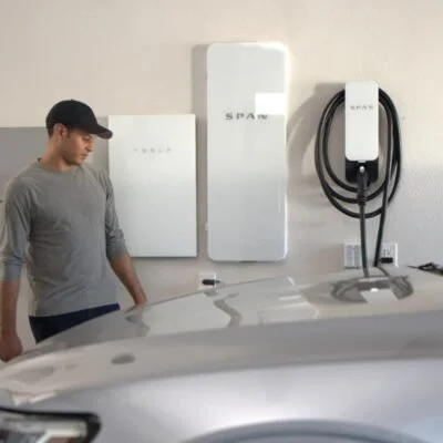 A man in a gray shirt and black cap standing next to an electric vehicle charging station on a white wall, with a charging unit and cable mounted on the wall.