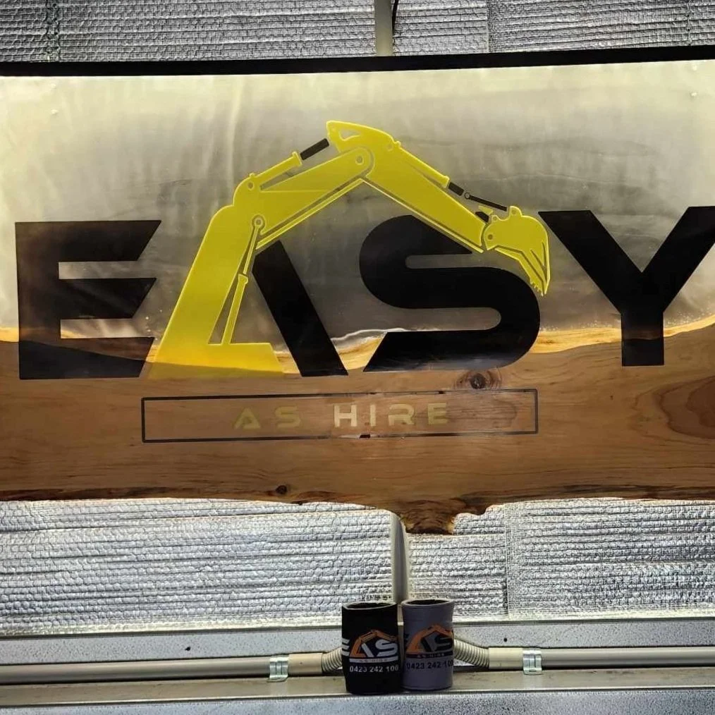 Sign for a business called 'EASY' featuring a yellow construction excavator as part of the logo. The sign includes the words 'AS HIRE' below the main text. The background shows a ceiling with metallic and textured surfaces.