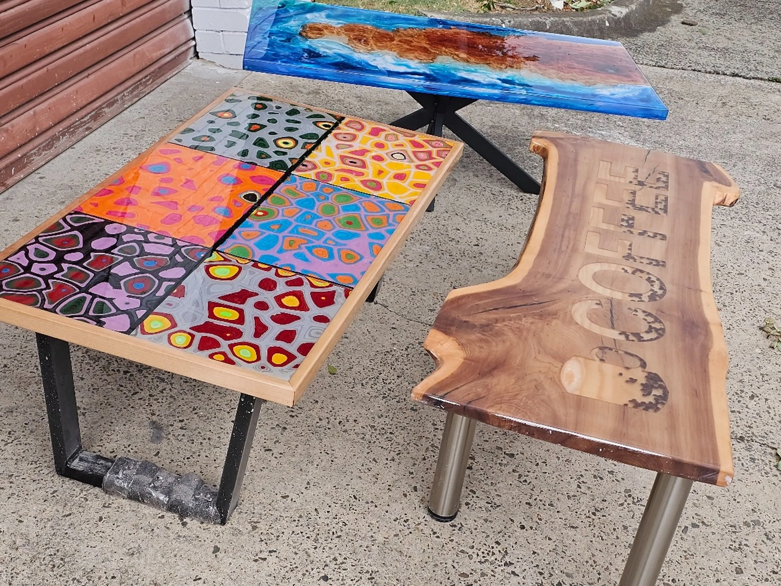 Three tables of different designs are placed outdoors on a concrete surface. The table on the left has a colorful, abstract, geometric pattern with various bright colors. The middle table features a vibrant, abstract rainbow pattern, and the table on the right is made of wood, has a natural finish, and the word 'COFFEE' carved into it.
