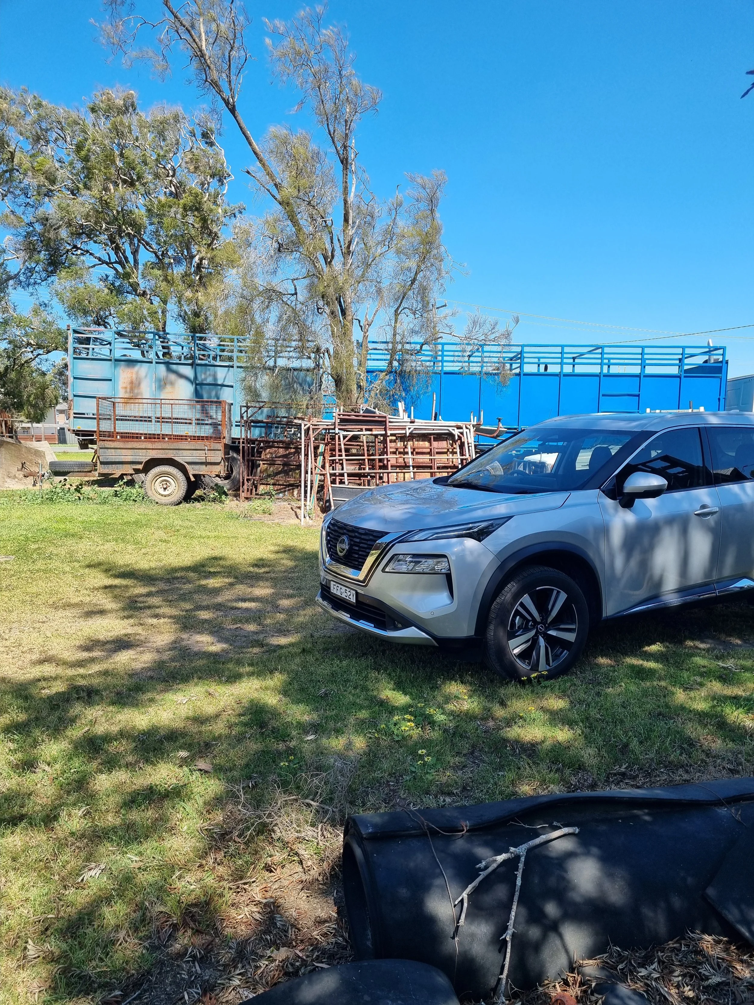 A silver car parked on a grassy area with trees and blue sky in the background. There are old trailers, metal frames, and a large blue structure or enclosure behind the car.
