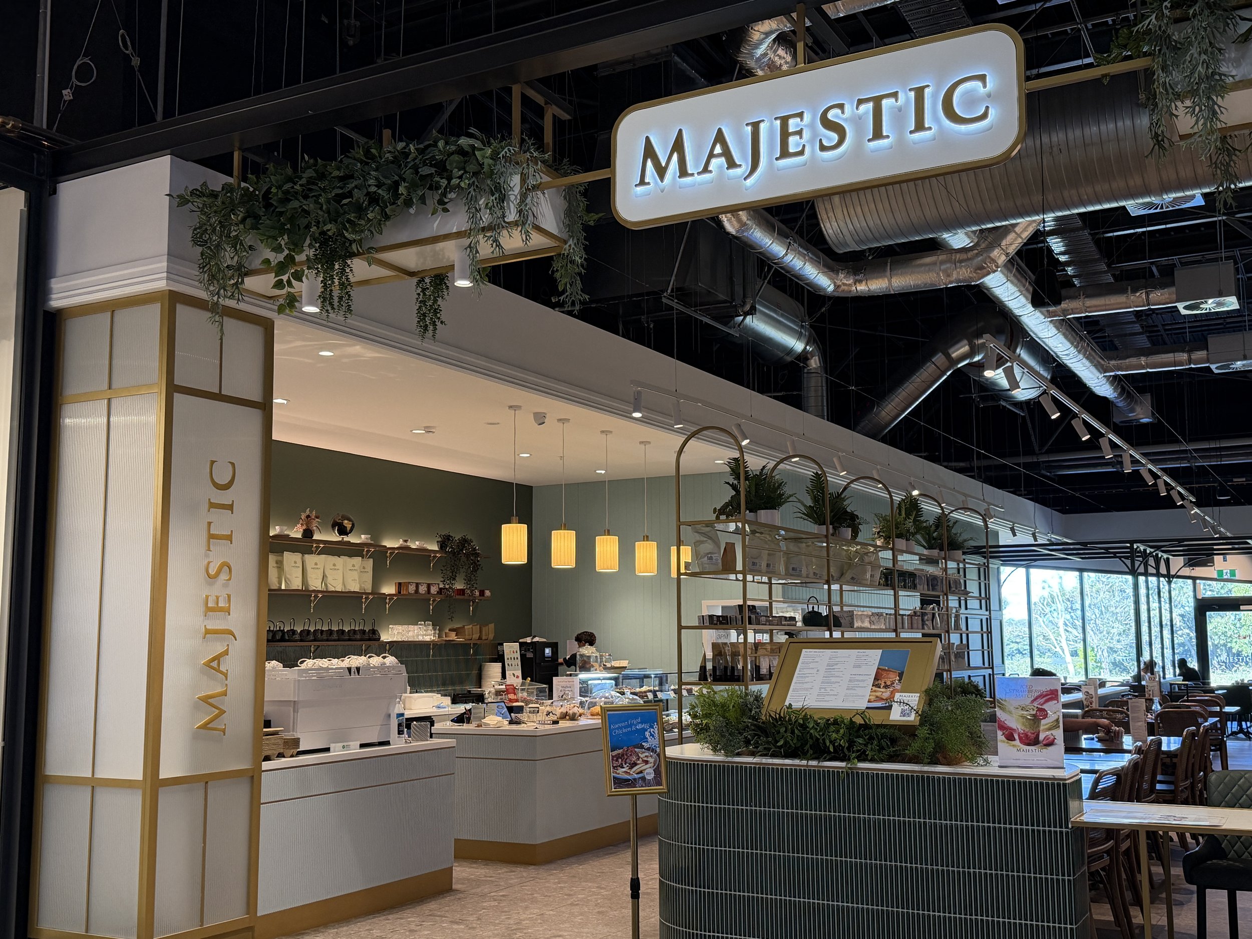 Interior of a shopping mall food court with a sign reading 'Majestic' in illuminated letters, greenery, and various food stands and seating areas visible.