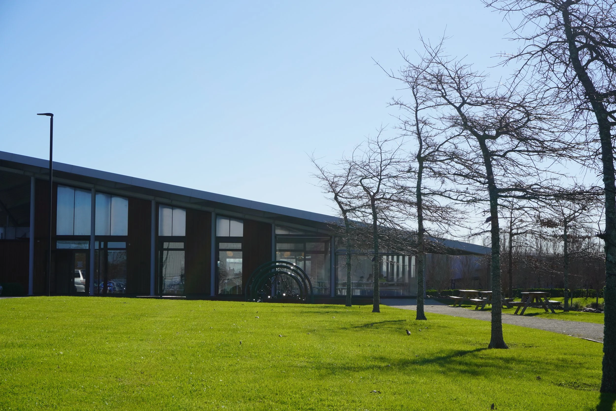 A modern building with large glass windows, surrounded by a well-maintained green lawn and several leafless trees, under a clear blue sky.