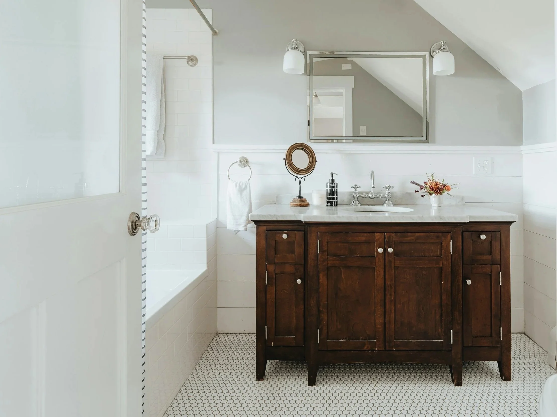 A renovated bathroom with a wooden vanity, white countertop, sink, mirror, and white tiled walls and floor in Saratoga Springs.