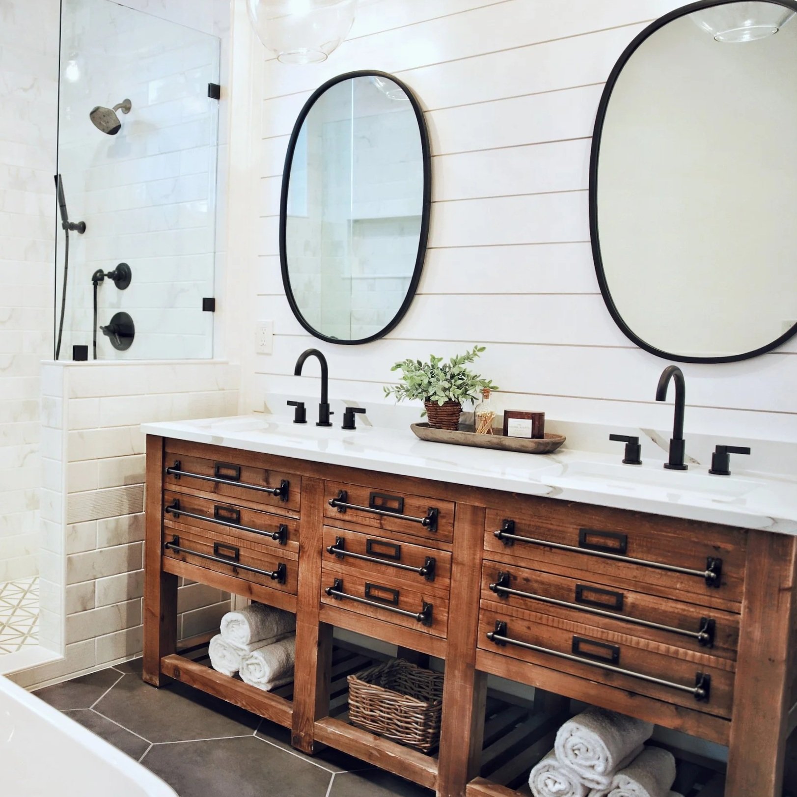 Custom primary bathroom renovation in the Lake George region featuring a custom wood double vanity, quartz countertops, frameless glass shower, freestanding soaking tub, large format tile flooring, undermount vanity sinks, and black faucets..