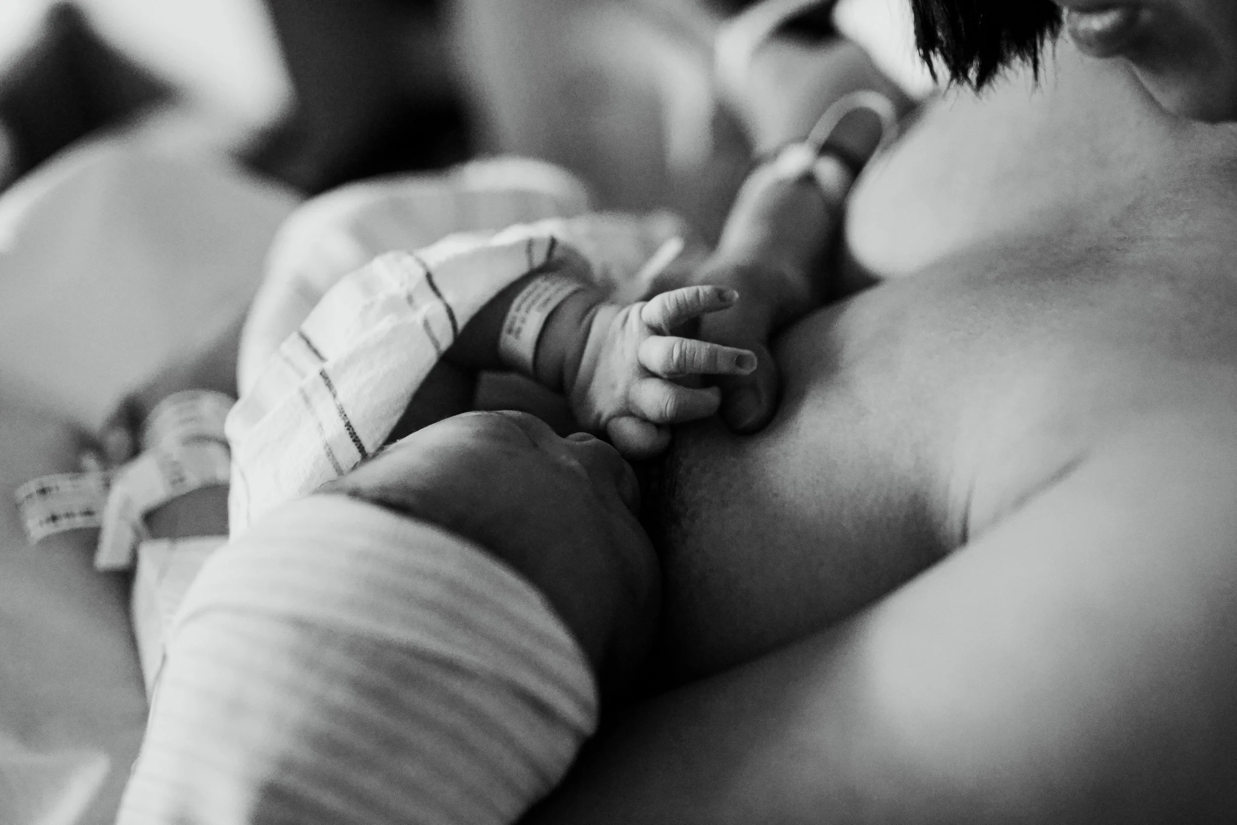 A black and white photo of a newborn baby breastfeeding, held by a woman, with a hospital wristband visible on the baby's wrist.