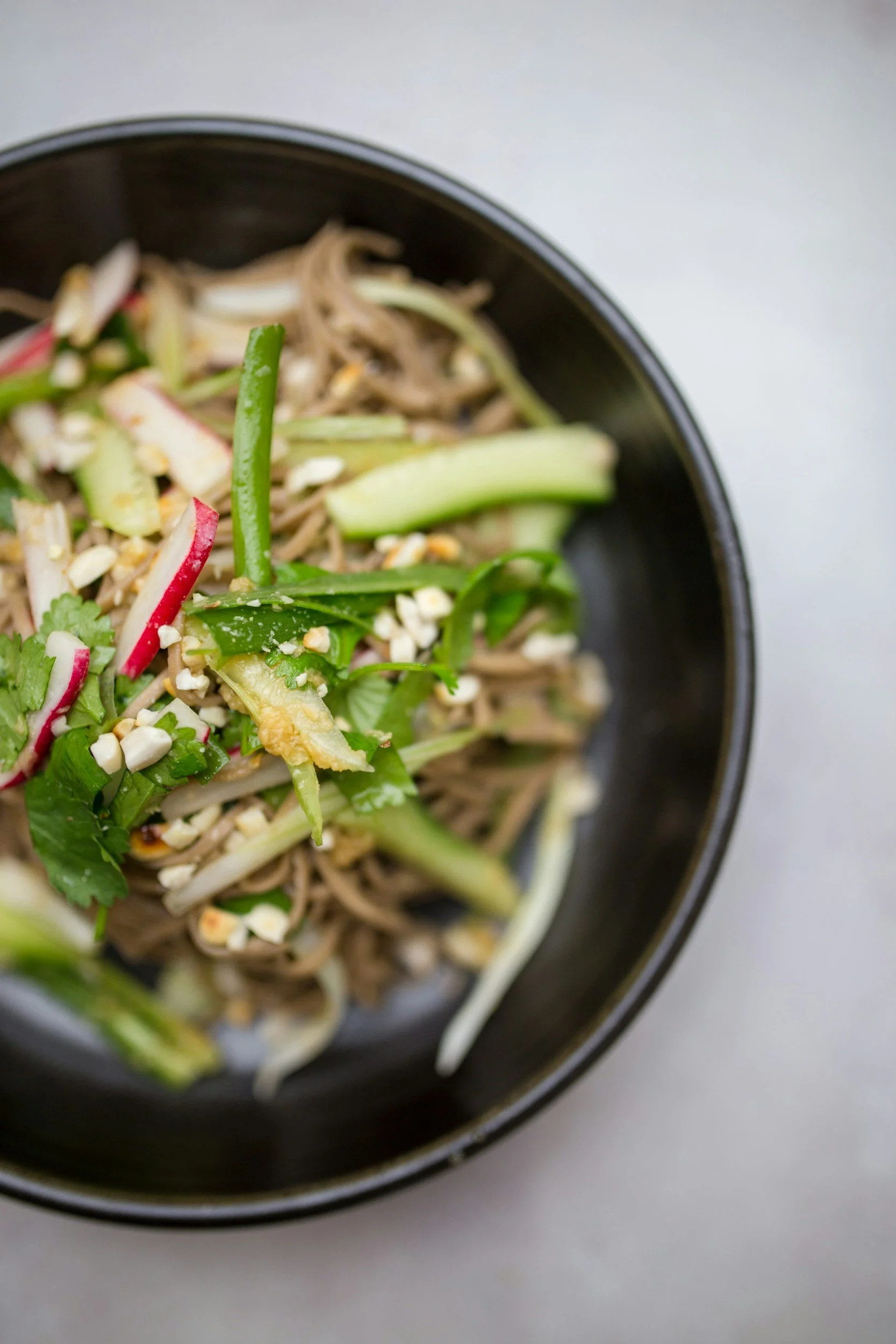 Salad with shredded carrots, cucumber, radish, cilantro, bean sprouts, chopped nuts, and sliced green onions in a dark bowl on a light surface.