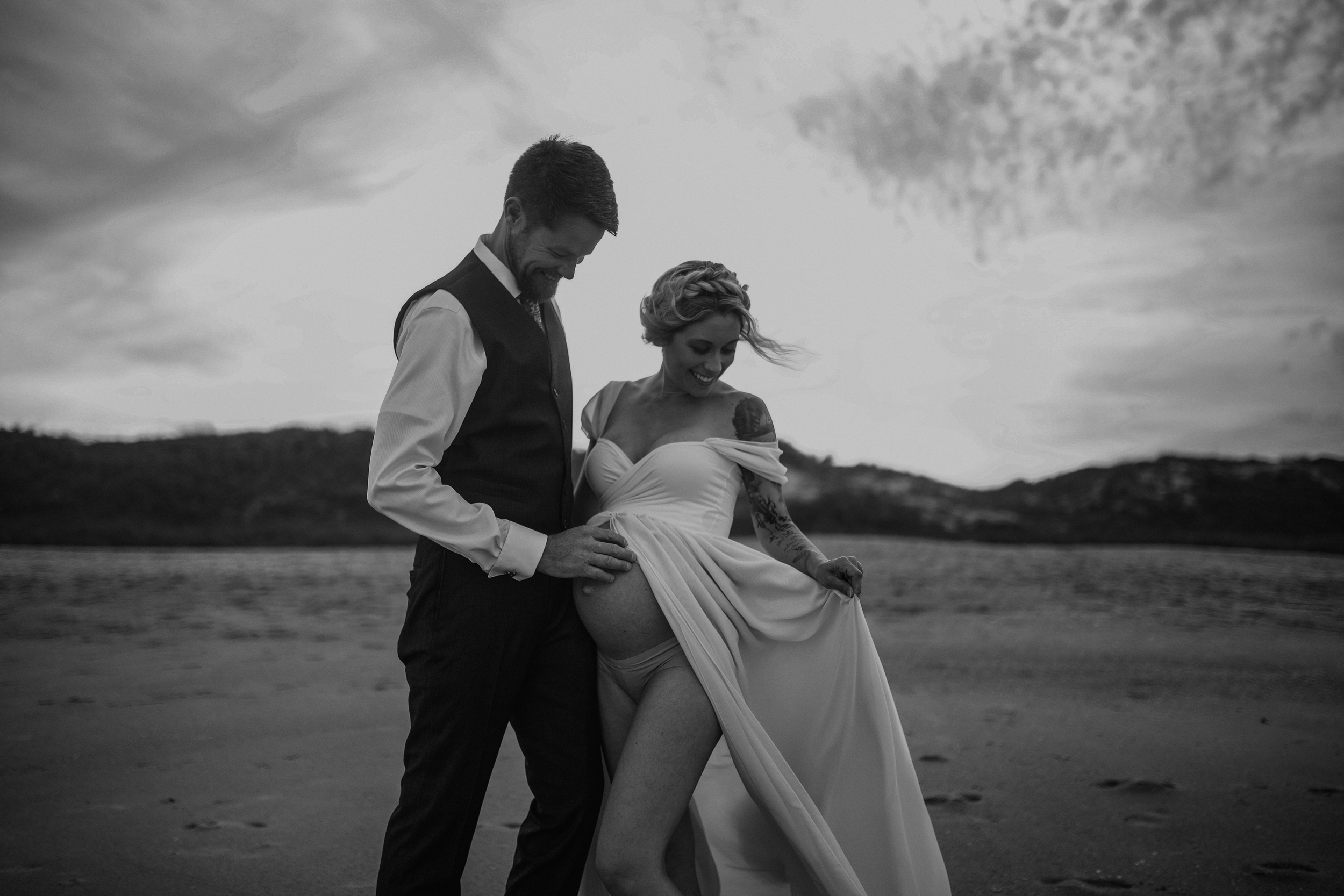 A man and woman in wedding attire standing on beach, smiling and holding her dress up to reveal her leg, with mountains and cloudy sky in background.