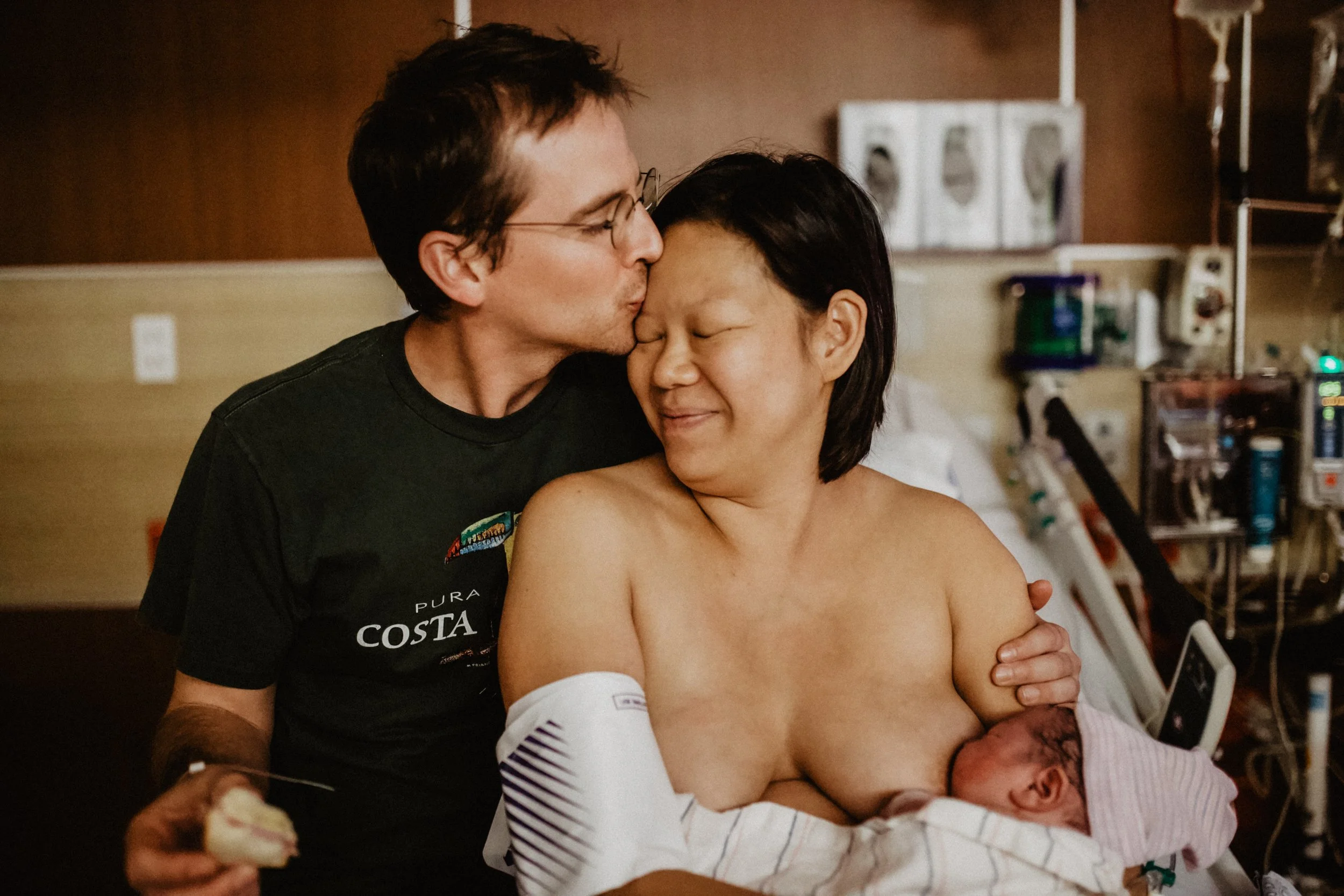 A man kisses a woman on the forehead in a hospital room. The woman is holding a newborn baby wrapped in a blanket. Medical equipment surrounds them.
