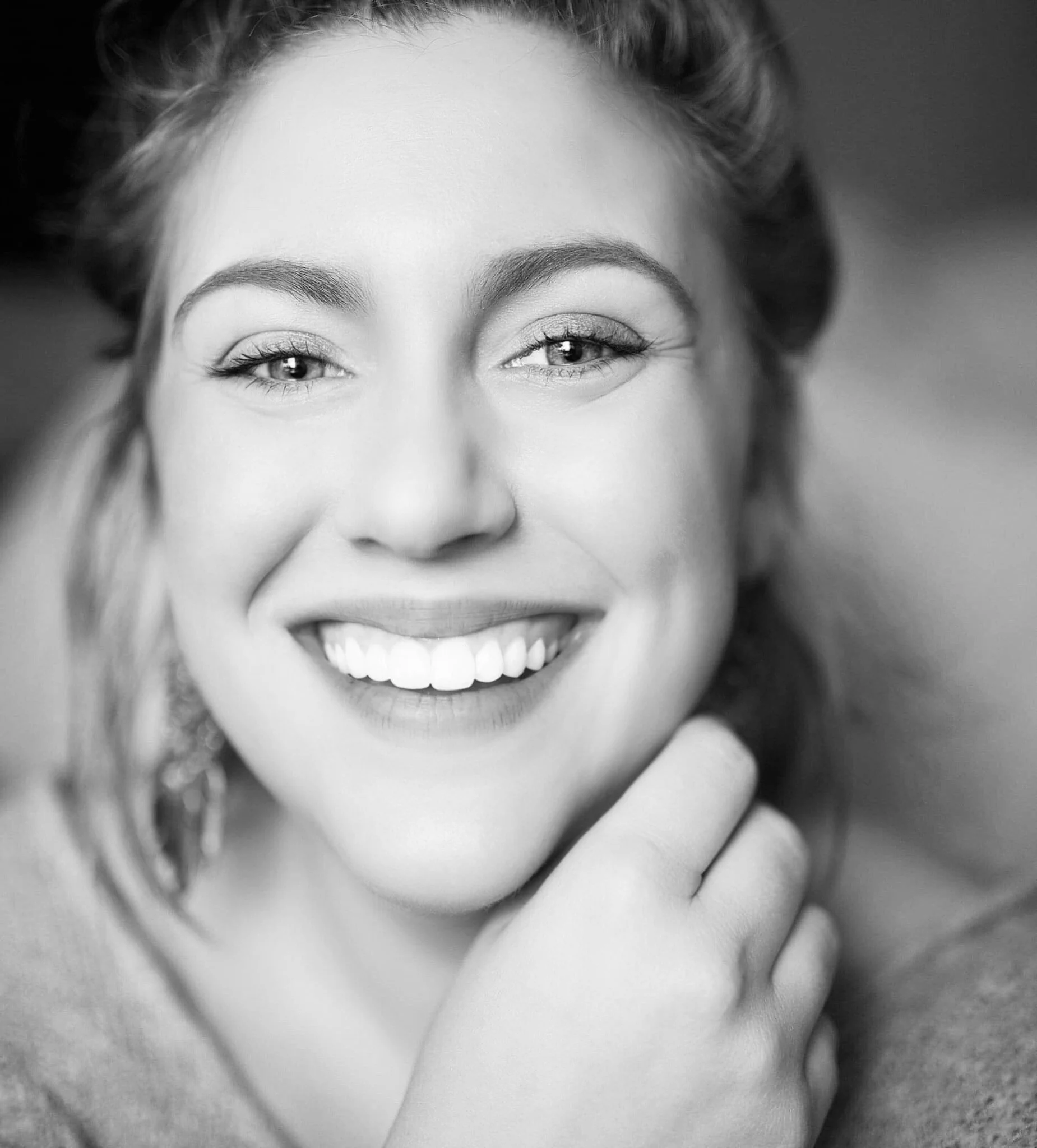 Black and white close-up portrait of a woman smiling, showing her teeth, with her hand resting near her chin.