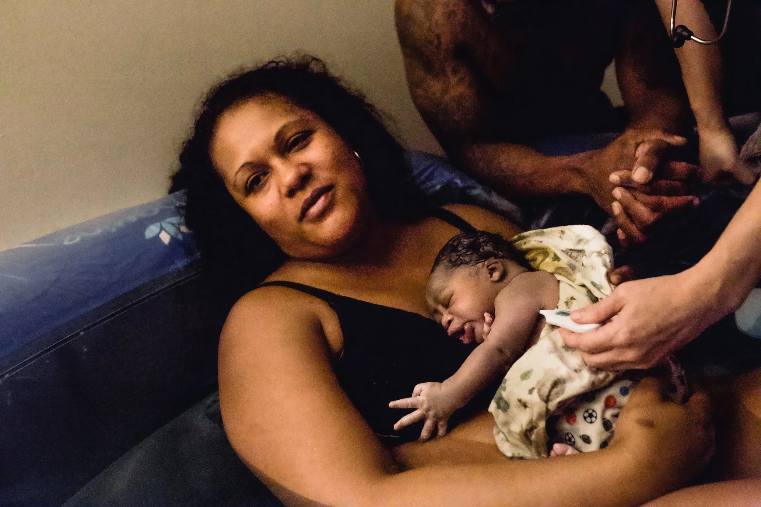 A woman lying on a bed holding a newborn baby, with a man sitting beside her clasping his hands.