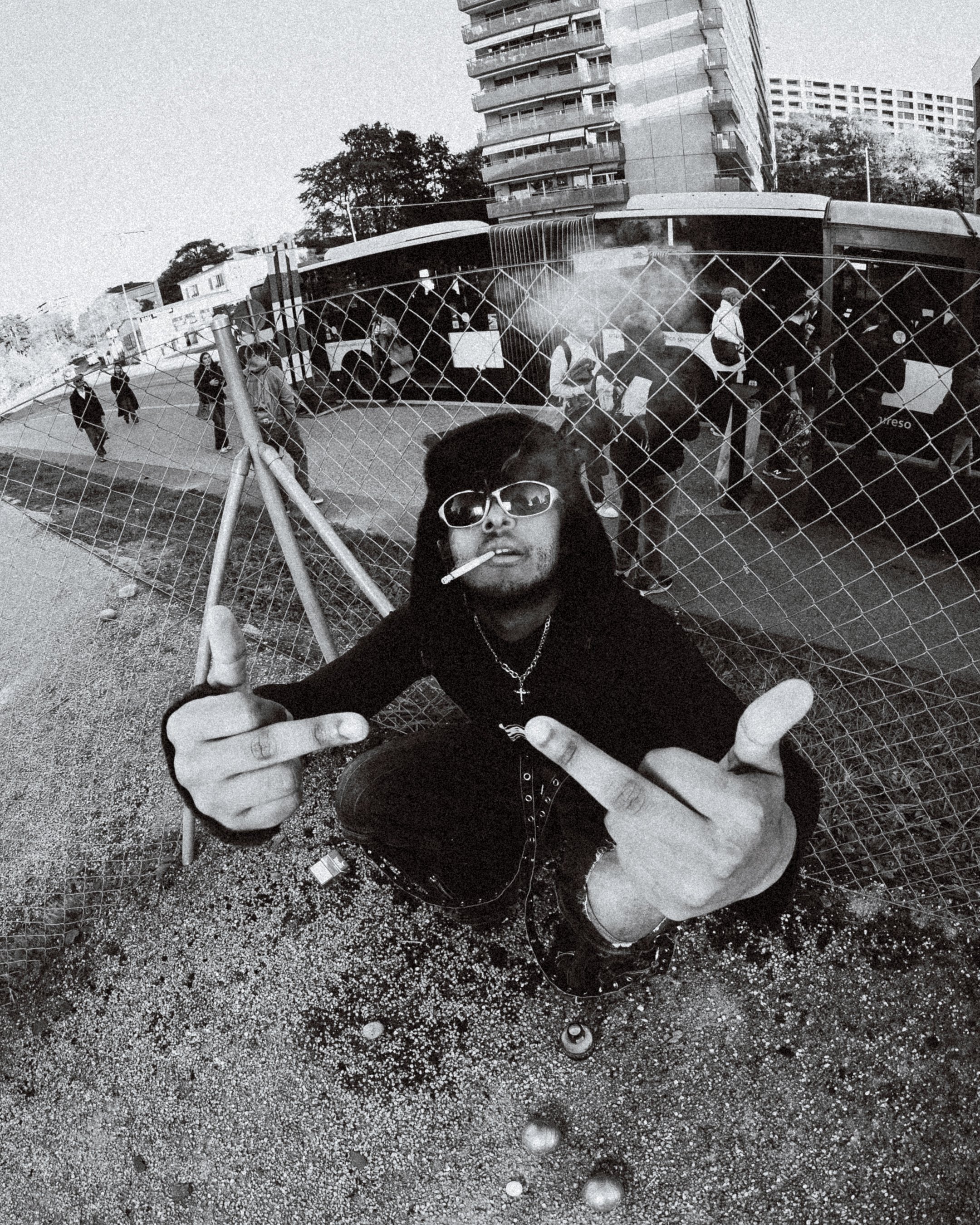 Un jeune homme portant des lunettes de soleil et un collier croix, faisant un signe avec ses mains, posé devant une clôture de chantier avec un fond urbain. En arrière-plan, des personnes entrent ou sortent d'un bus, avec des bâtiments modernes en arrière-plan.
