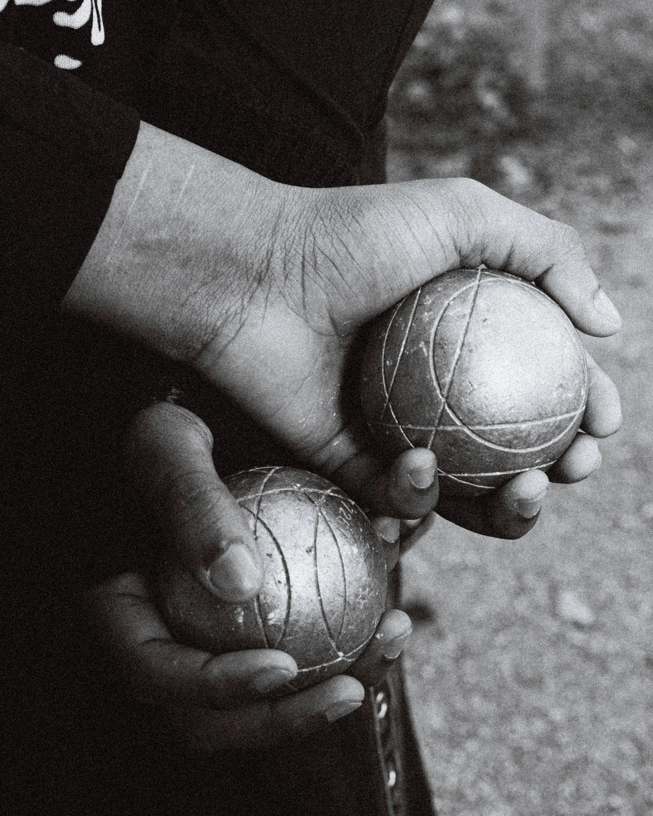 Deux mains d'enfant tenant deux balles de pétanque, une main adulte tenant une boule de pétanque, en noir et blanc.
