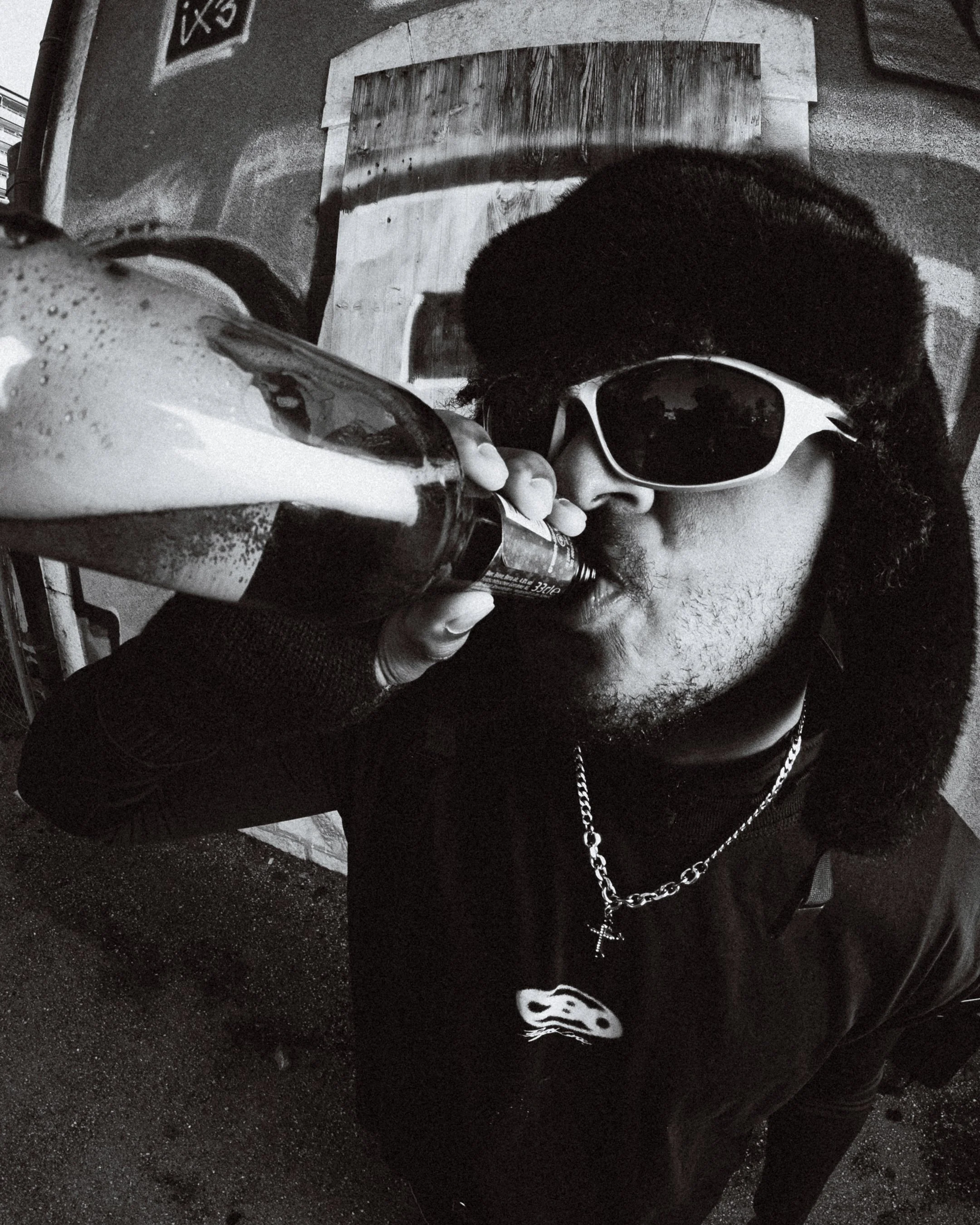 Un homme portant des lunettes de soleil blanches et un bonnet noir, habillé en noir, qui boit une boisson d'une bouteille.