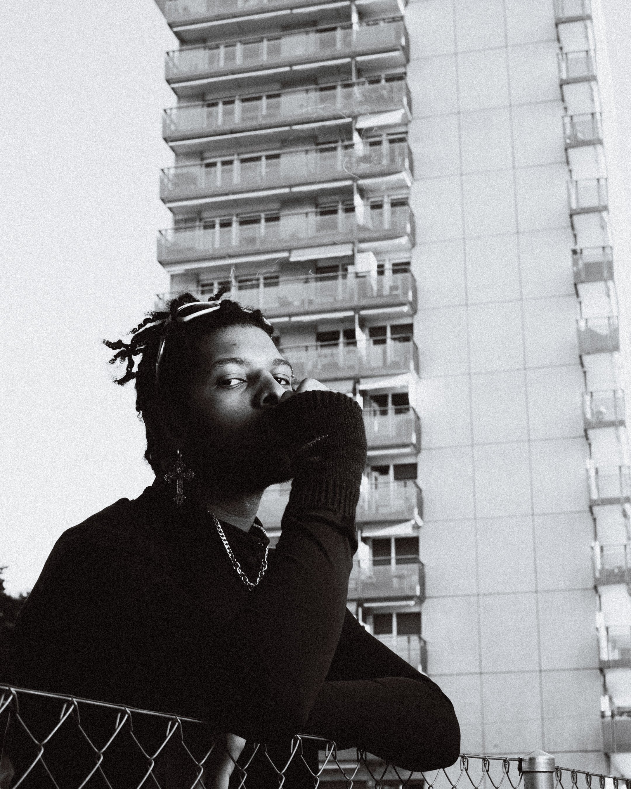 Une personne portant un manteau noir et un collier en chaîne, avec un seul gant noir, se tient à côté d'une clôture en métal devant un grand immeuble d'appartements. La photo est en noir et blanc.