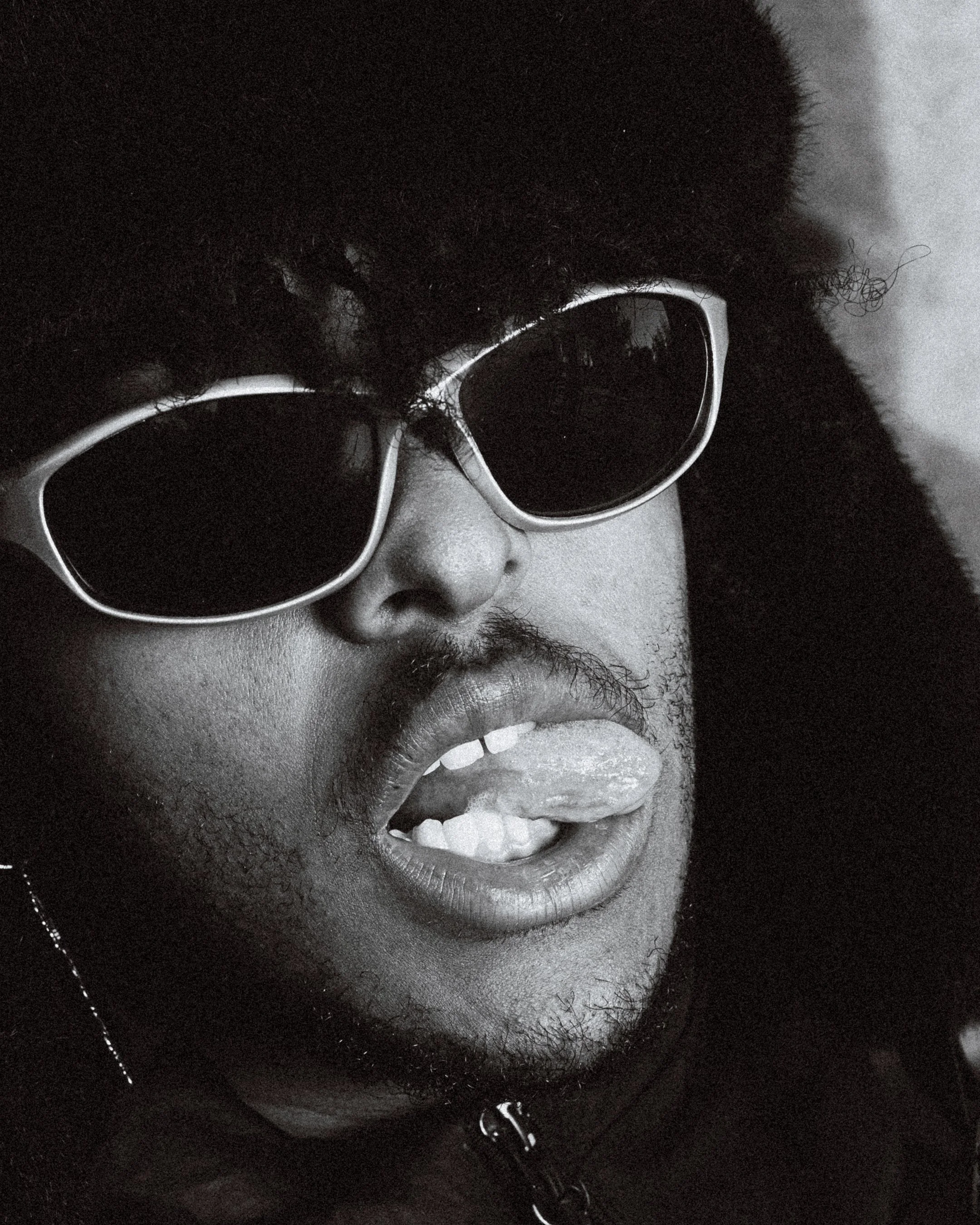 Portrait en noir et blanc d'une personne portant des lunettes de soleil, avec les lèvres ouvertes et la langue sortie.