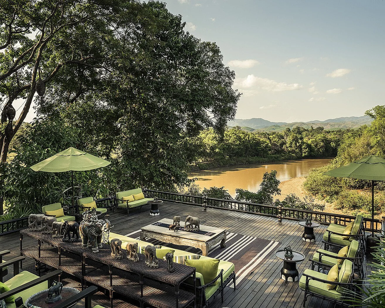 Open-air wooden terrace of a luxury jungle lodge with green cushioned seating, elephant sculptures and a rustic coffee table overlooking a wide river winding through dense forest, with misty mountains in the distance in northern Thailand