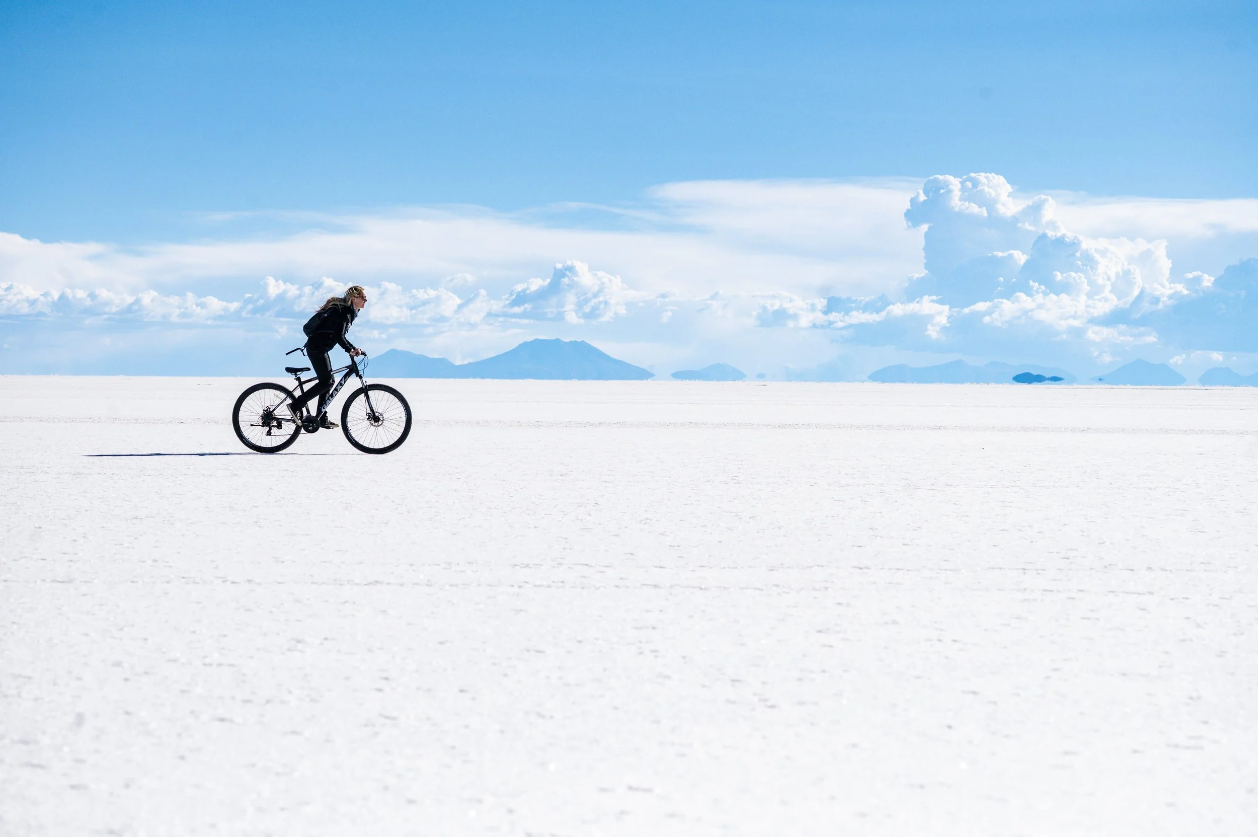 Traveller cycling alone across the blinding white surface of the Salar de Uyuni under a deep blue sky with mountains in the distance, Bolivia