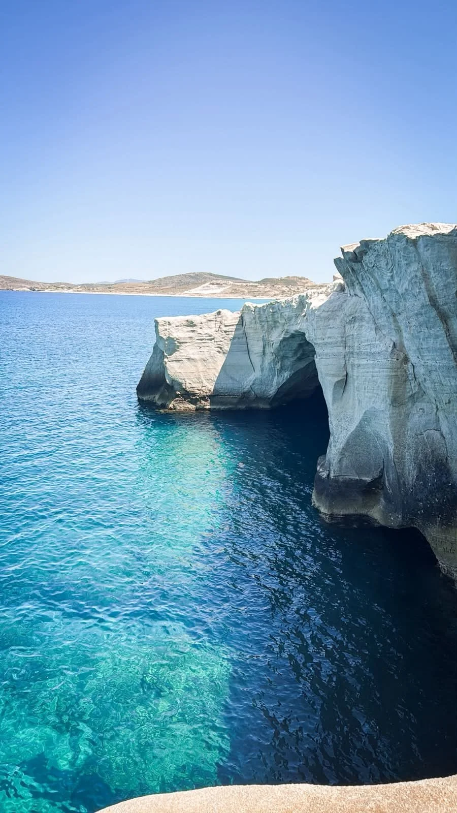 Dramatic white volcanic rock arches and cliffs rising from deep turquoise water along the coastline of Milos island, one of Greece's most visually striking Cycladic islands
