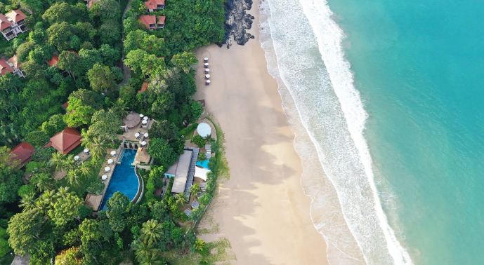 Aerial view of a tropical beach resort with a sandy shoreline, turquoise ocean waves, lush green trees, and outdoor amenities including pools and sun loungers.