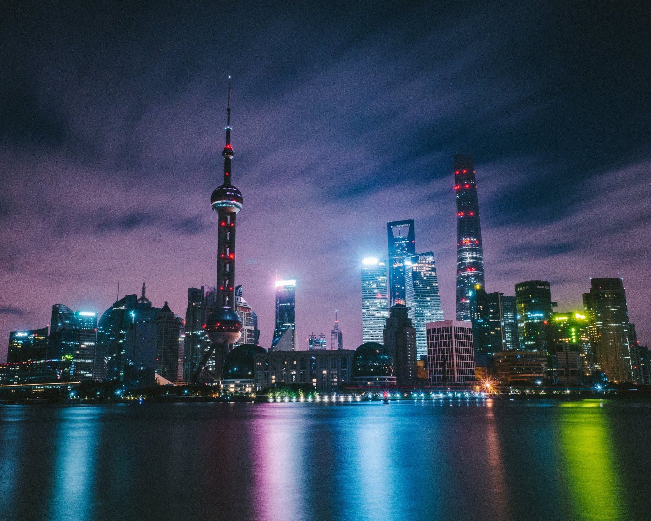 Shanghai's Pudong financial district skyline at night reflected in the Huangpu River, with the illuminated Oriental Pearl Tower, Shanghai Tower and World Financial Center glowing under a dramatic purple sky