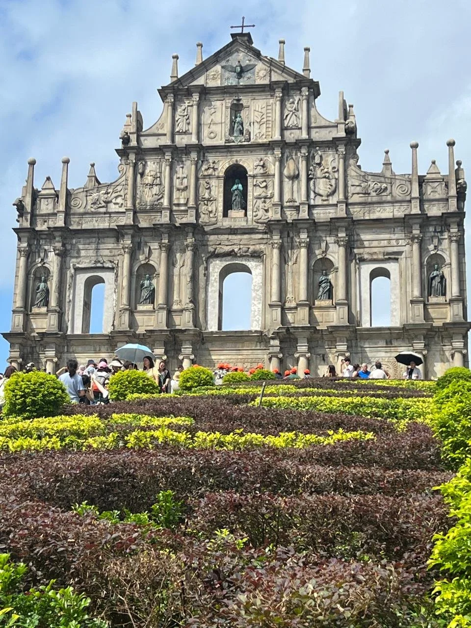 The ornate baroque stone facade of the Ruins of St Paul's Cathedral in Macau, a UNESCO World Heritage Site, with its carved tiers and statues standing against a blue sky, visitors visible below