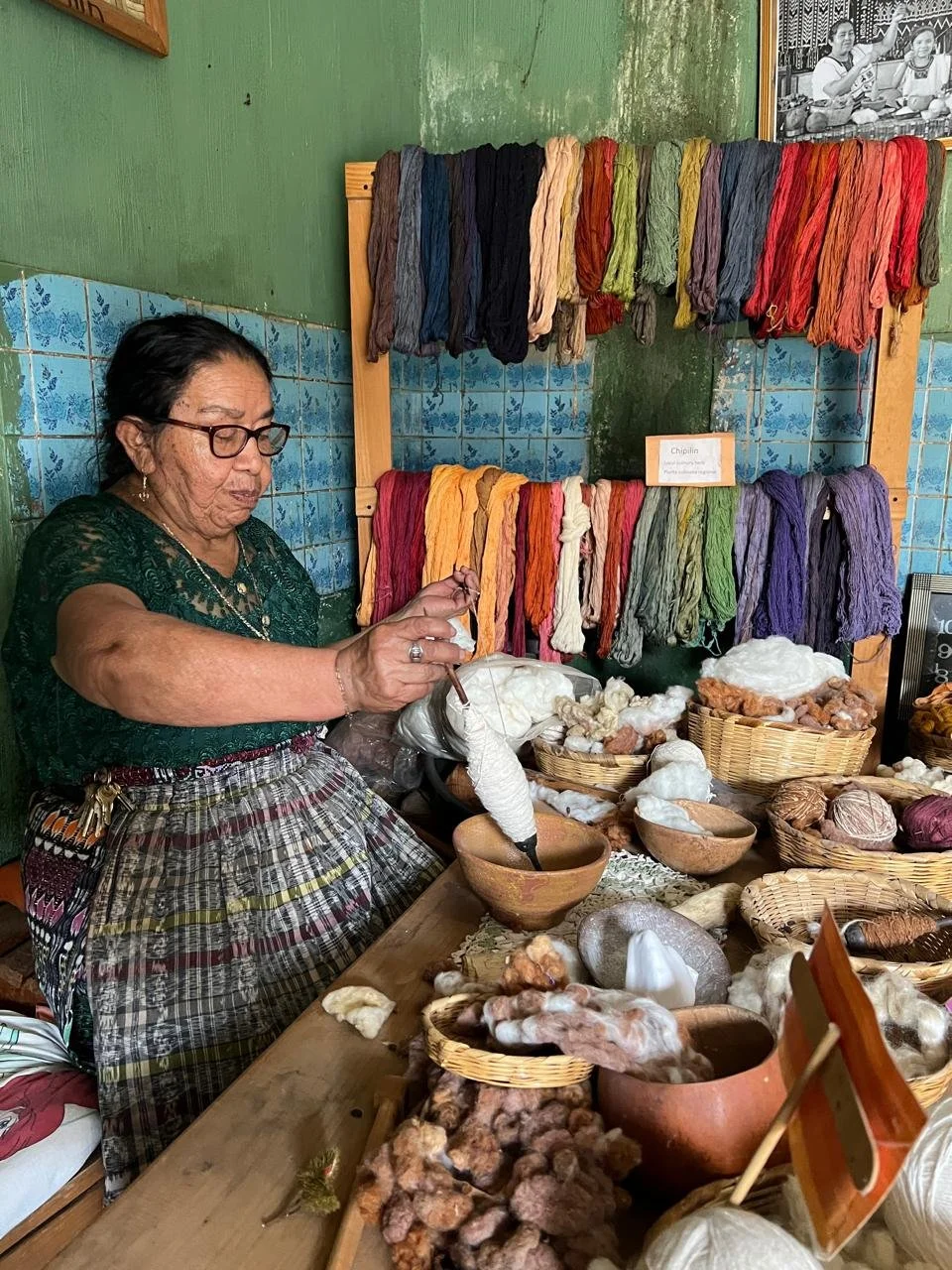 Indigenous Maya woman weaving traditional textiles surrounded by colourful woven goods and ceramics in a market stall near Lake Atitlán, Guatemala