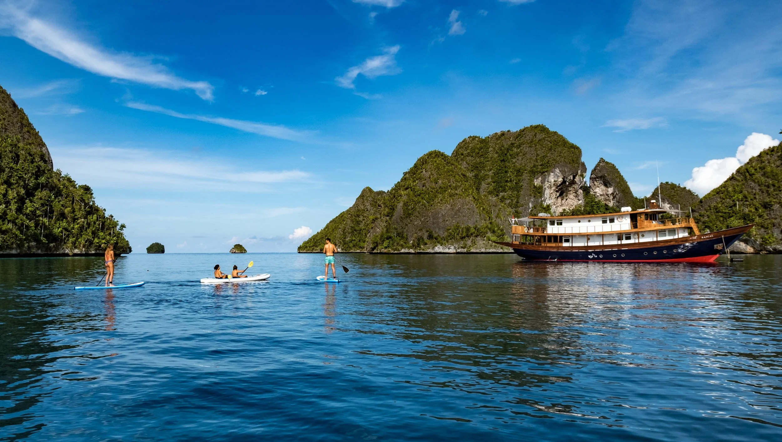 Guests paddleboarding beside a private liveaboard vessel anchored in calm blue waters among forested limestone islands in the Maluku archipelago, Indonesia