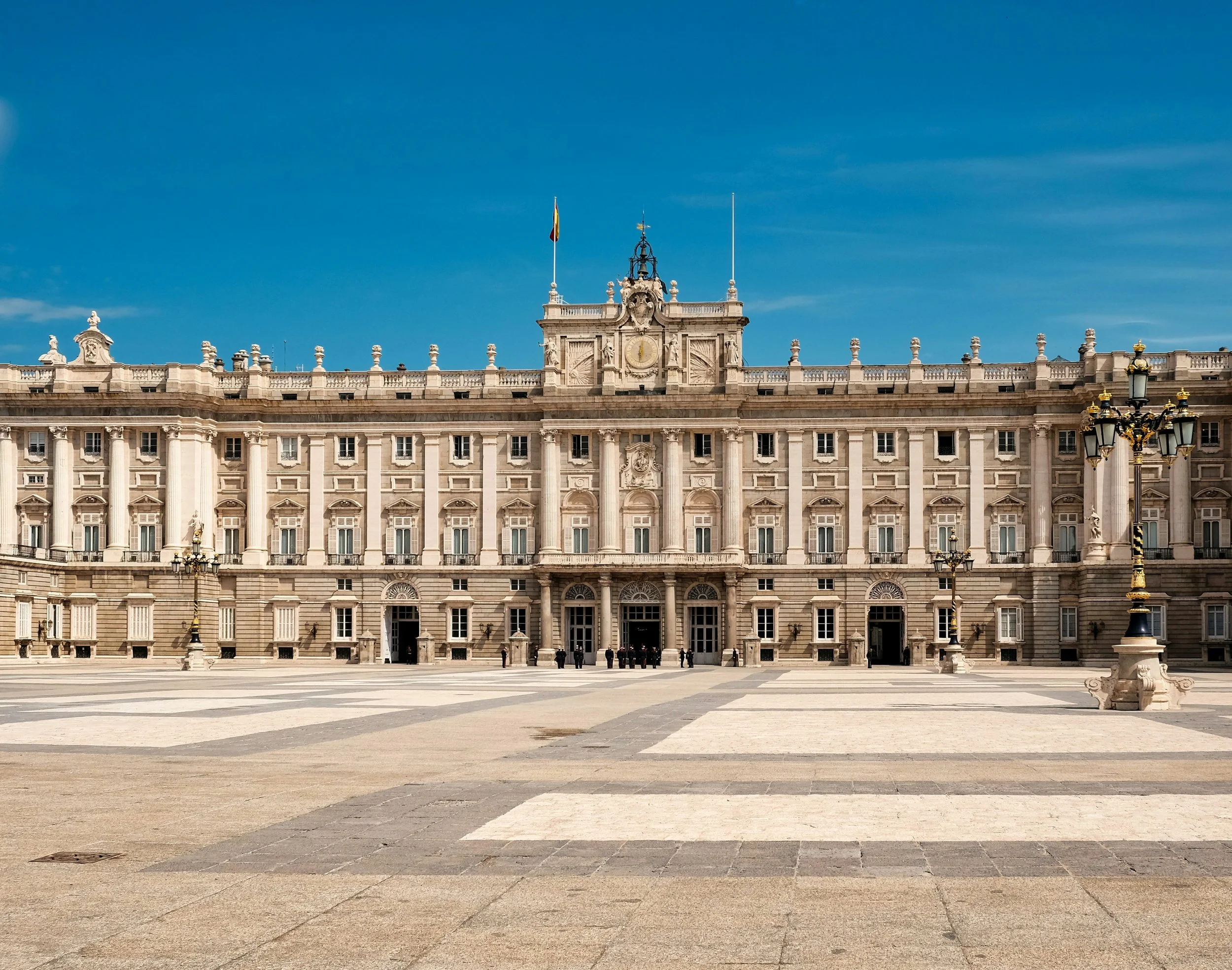 The neoclassical limestone façade of the Royal Palace of Madrid under a clear blue sky, with guards at the entrance and ornate lamp posts in the forecourt, Spain