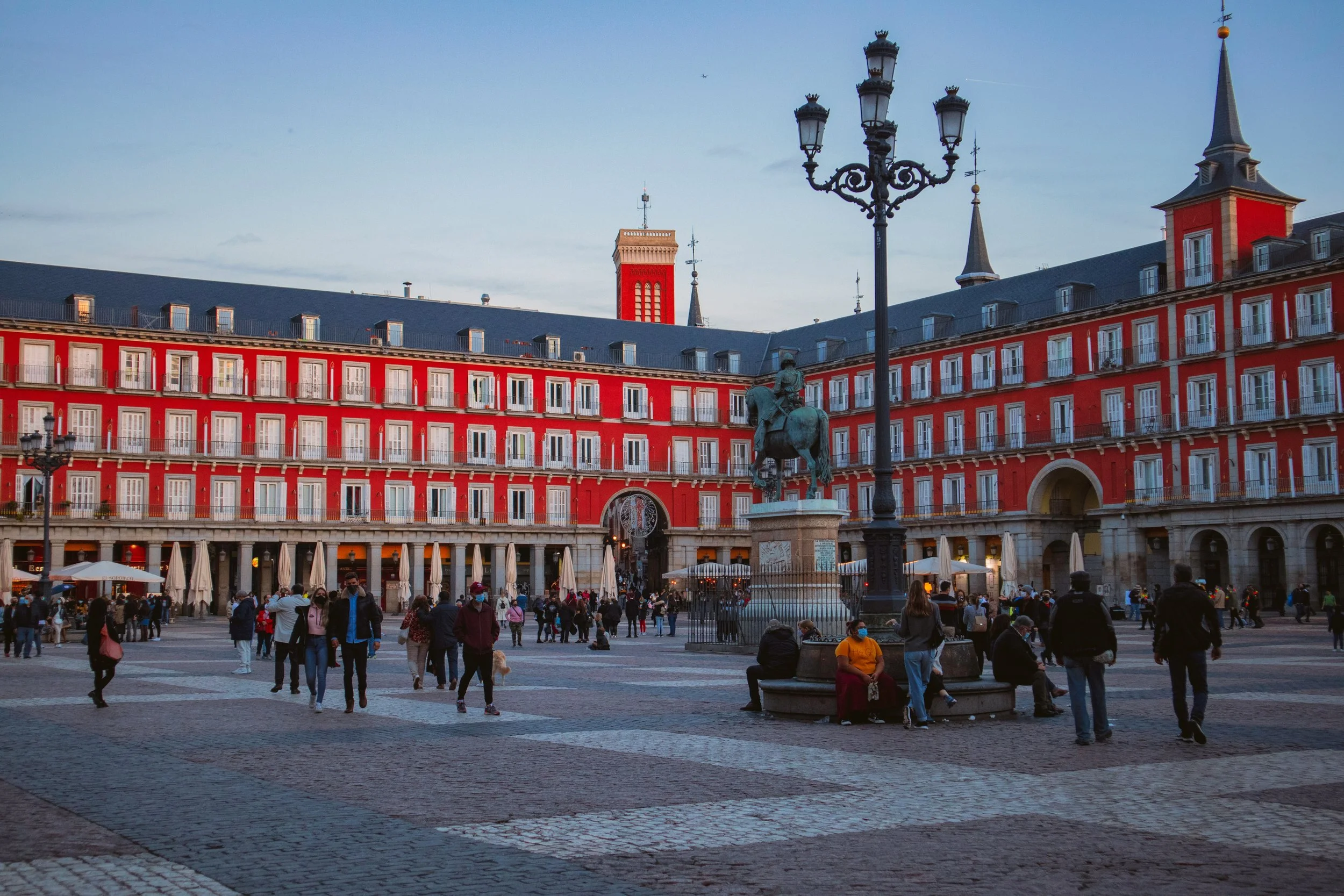 Plaza Mayor in Madrid at dusk, with its iconic red-painted Habsburg façades, the equestrian statue of King Philip III and crowds of locals gathered in the cobblestone square, Spain