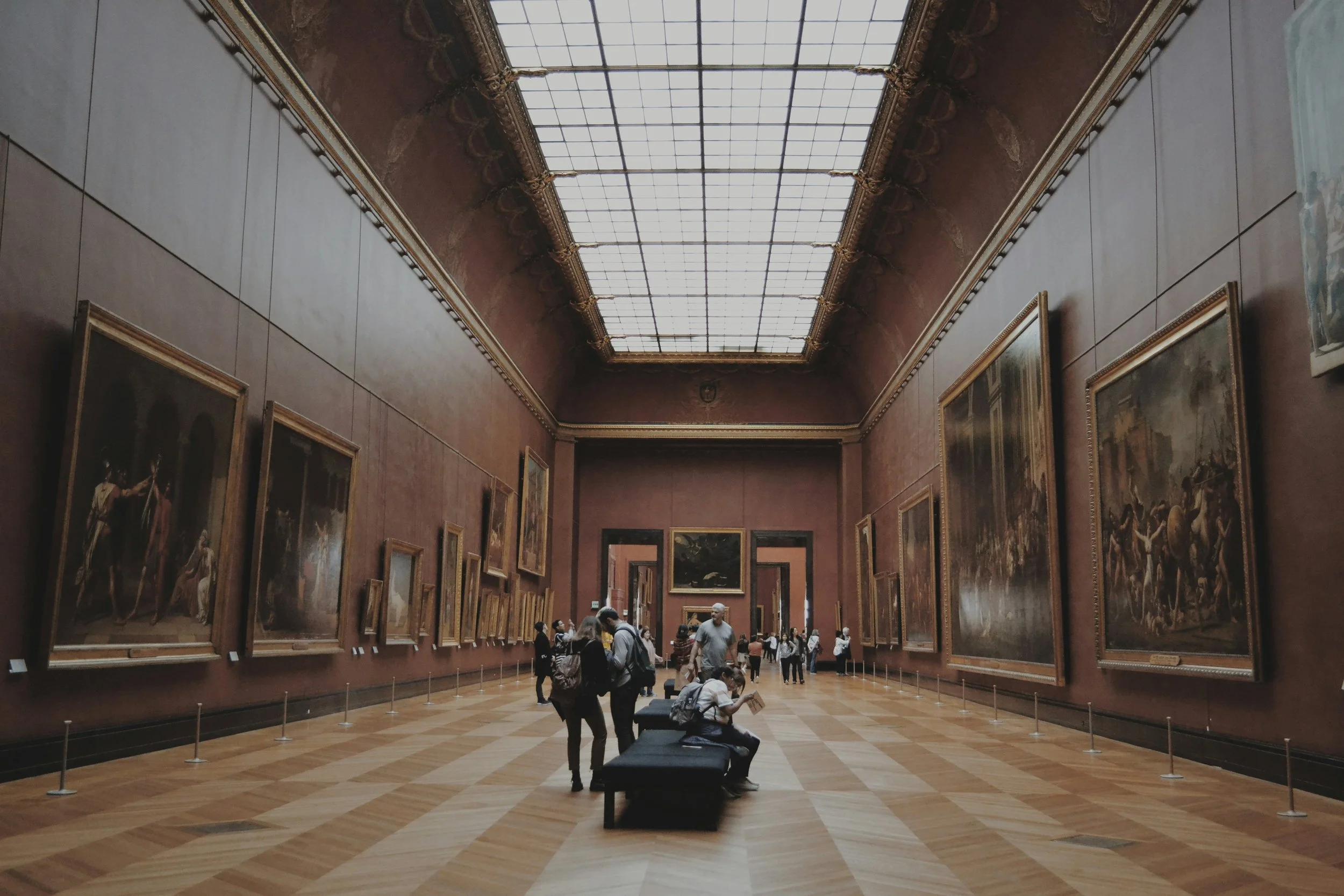 A long grand gallery hall in the Prado Museum in Madrid, lined with large Old Master paintings on deep red walls beneath a skylit ceiling, with visitors studying the works, Spain
