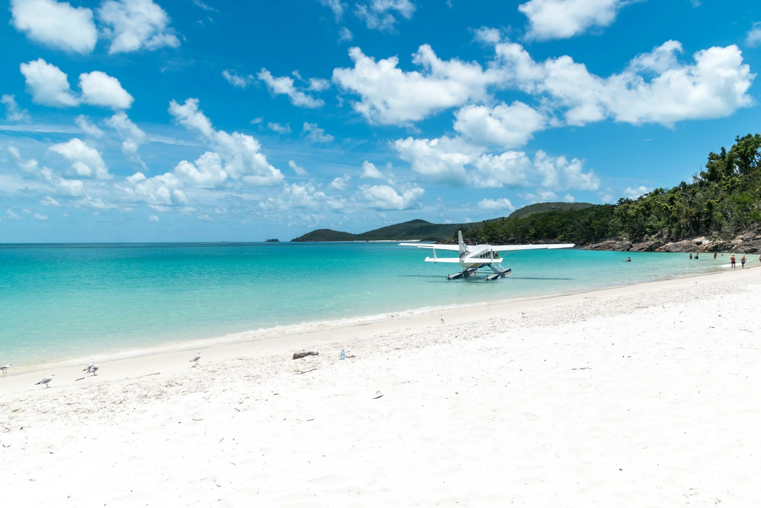 A sailboat floating in a narrow, turquoise bay between green, rocky hills on a bright, sunny day.