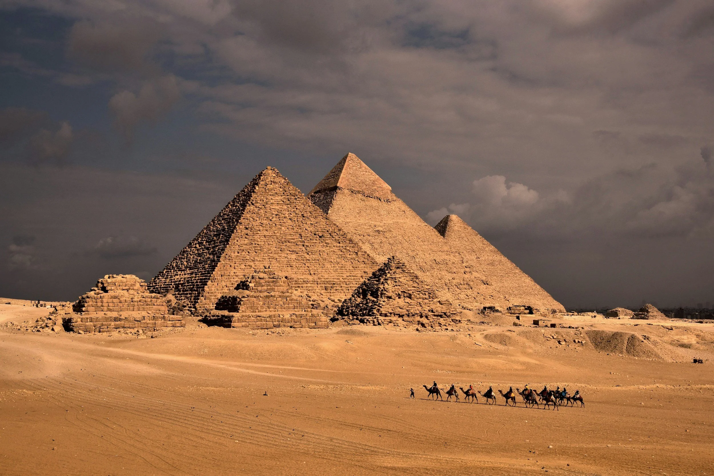 The Great Pyramid of Giza with camels and riders in the desert in Egypt under a cloudy sky.