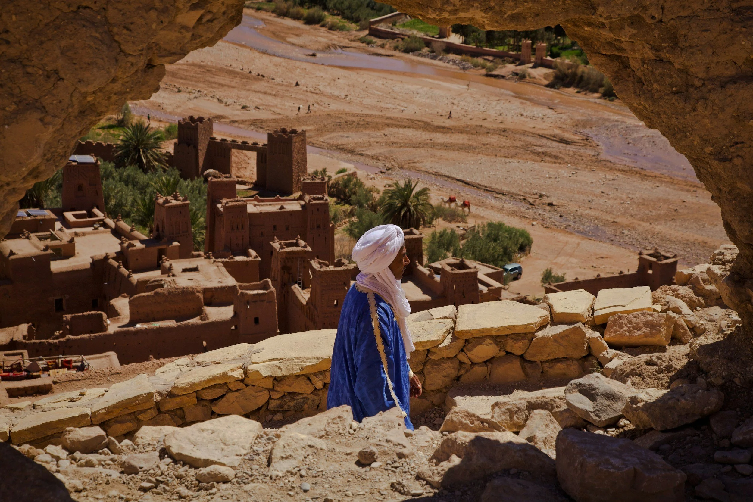 A person dressed in traditional Moroccan clothing, wearing a white turban and blue robe, walking on a rocky path overlooking ancient mud-brick buildings in a desert town with palm trees and a river in the background.