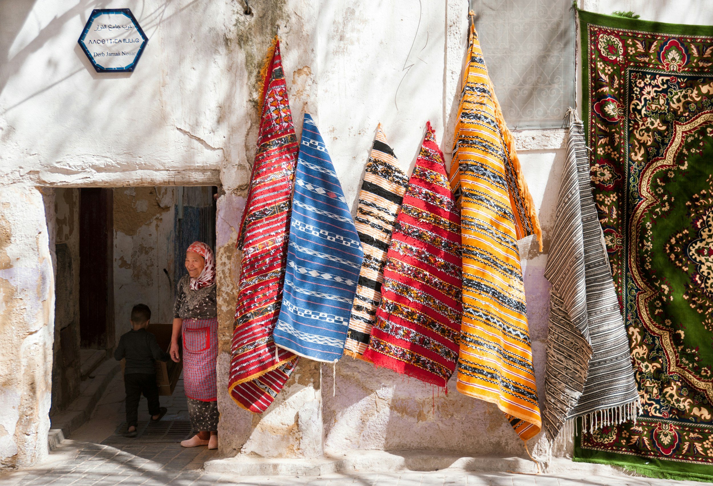Colorful traditional rugs hanging outside a building with a woman and a child nearby.
