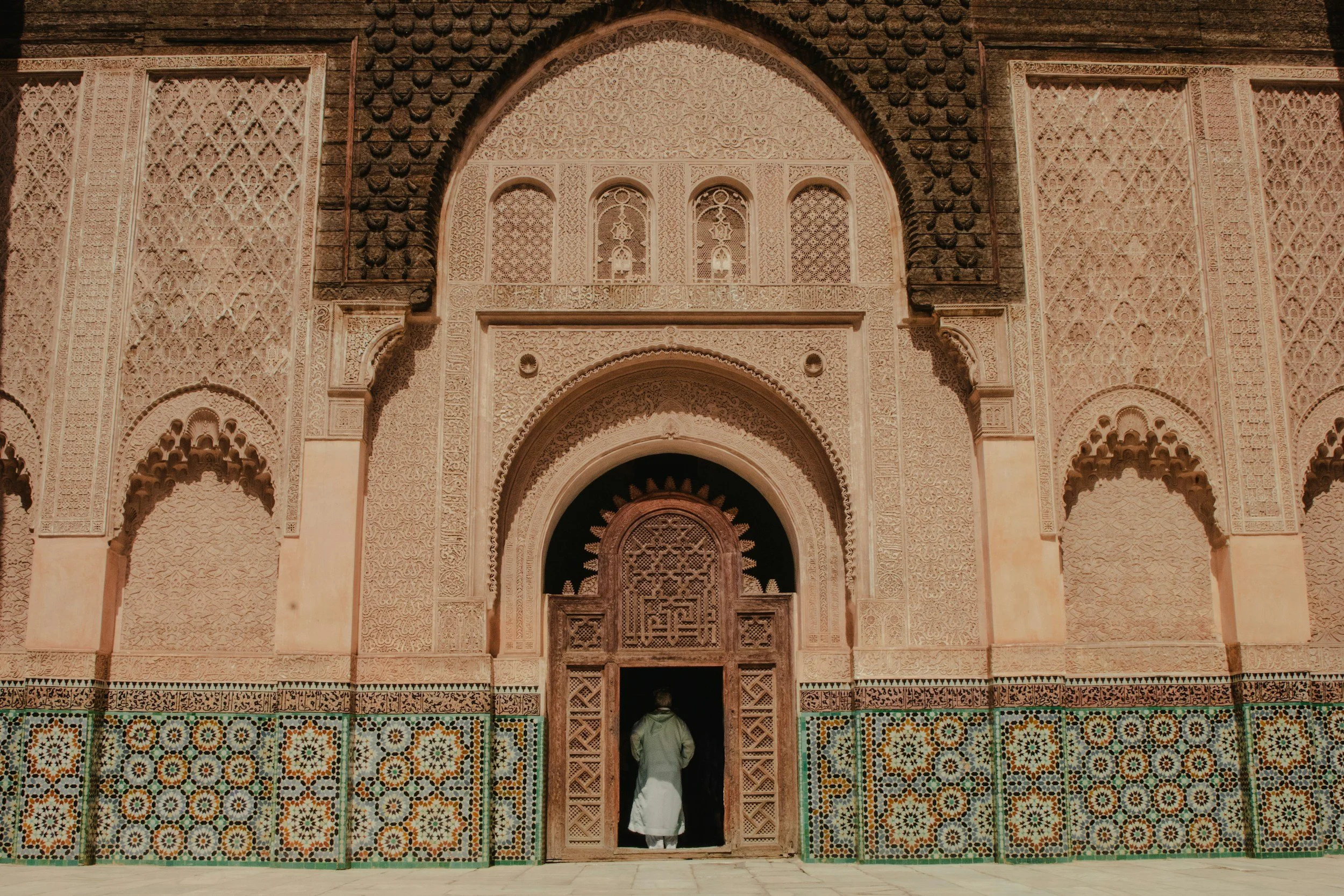 A person in traditional clothing walking into an intricately decorated mosque entrance with detailed geometric and floral patterns on the walls and tiled lower sections.