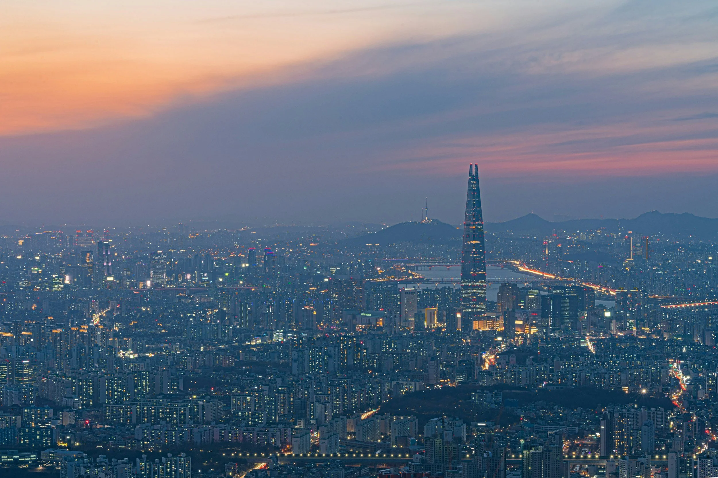 Panoramic aerial view of Seoul at twilight showing the illuminated cityscape, Lotte World Tower, Han River bridges, and Namsan mountain under a pink and purple sky