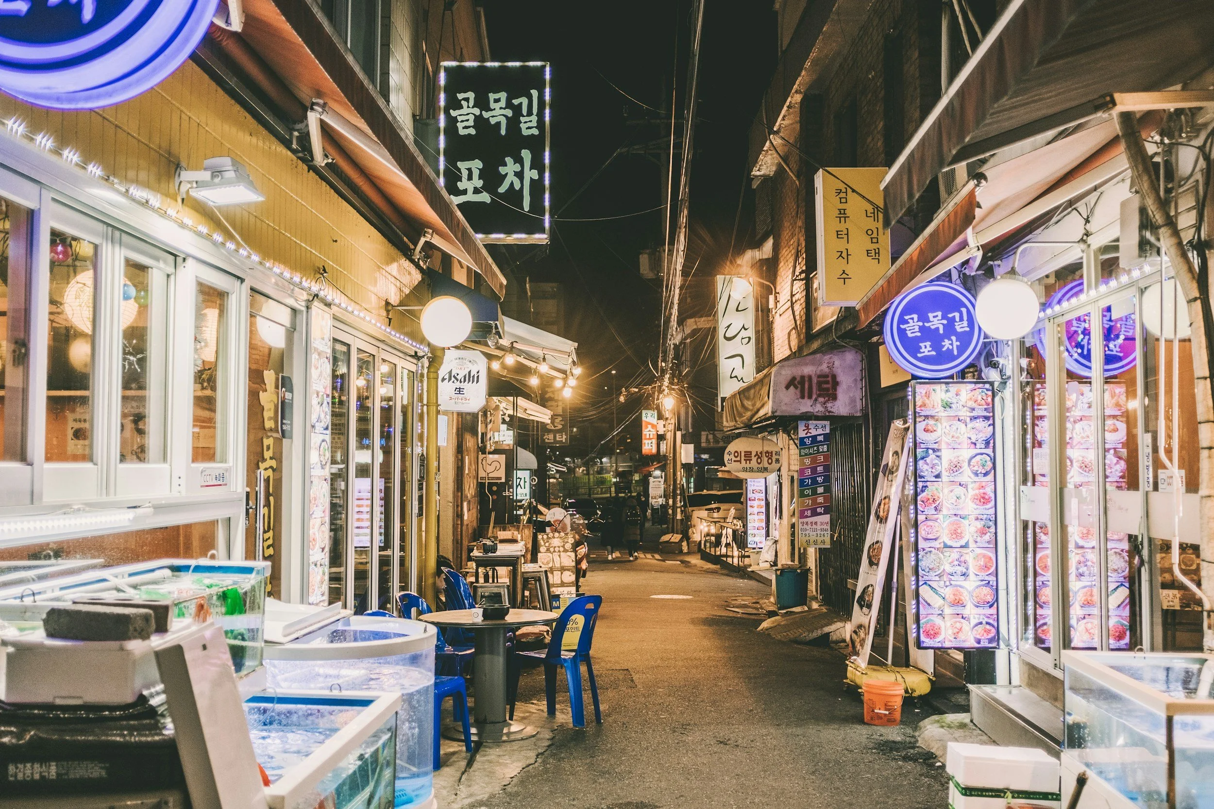 Narrow Seoul alley lit by neon signs and street lanterns at night, lined with pojangmacha food stalls and small restaurants with Korean signage