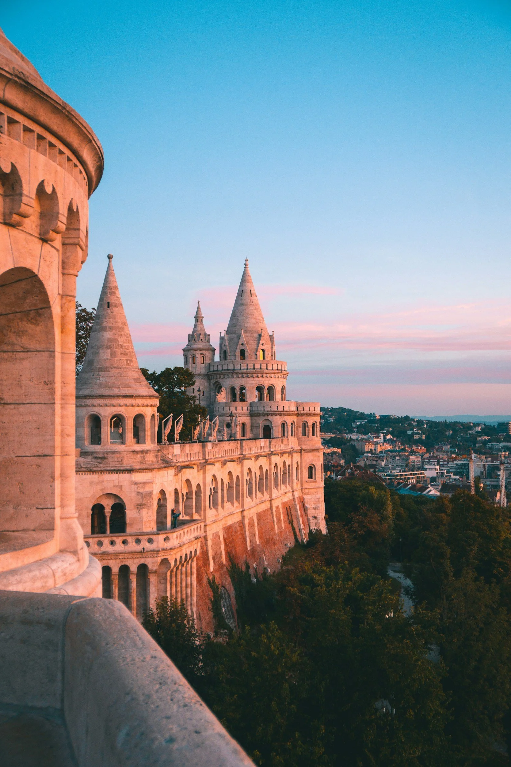 Neo-Romanesque stone towers and arcaded terraces of Fisherman's Bastion glowing pink at sunset on Castle Hill in Budapest, Hungary, with the city stretching into the distance below
